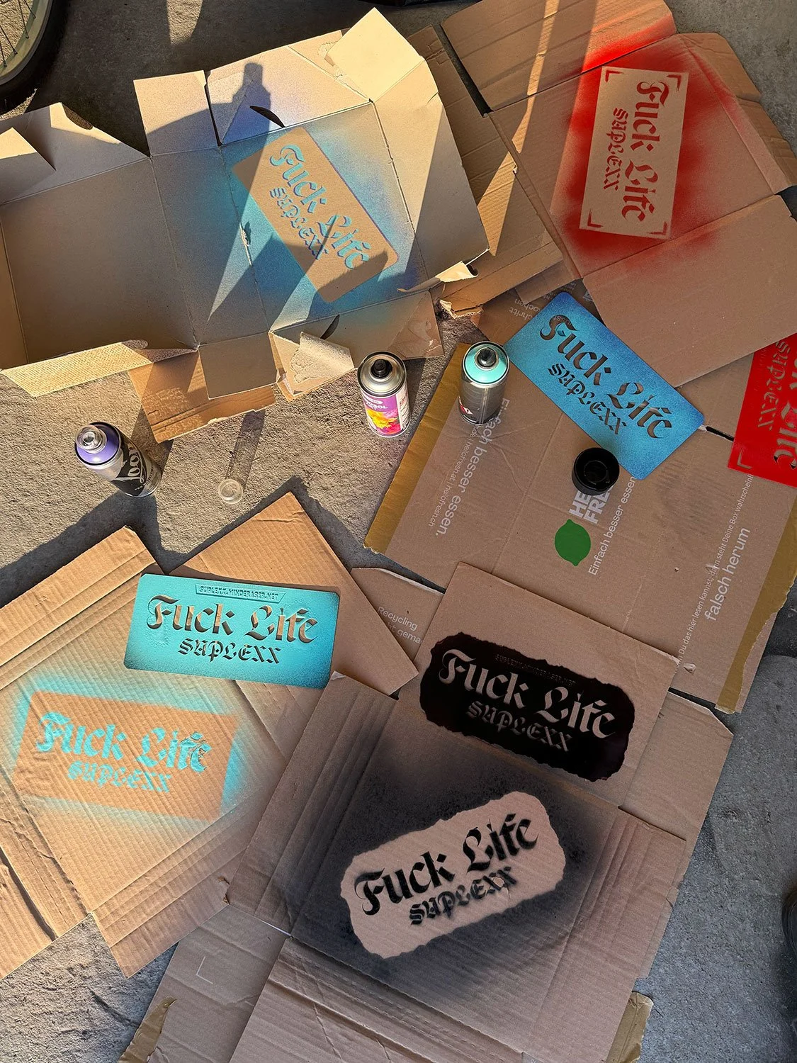 A collection of flattened cardboard boxes with spray-painted slogans and logos, including multiple examples of the phrase 'F*** Life' in various styles, placed on a concrete surface. There are also several spray paint cans around the boxes.