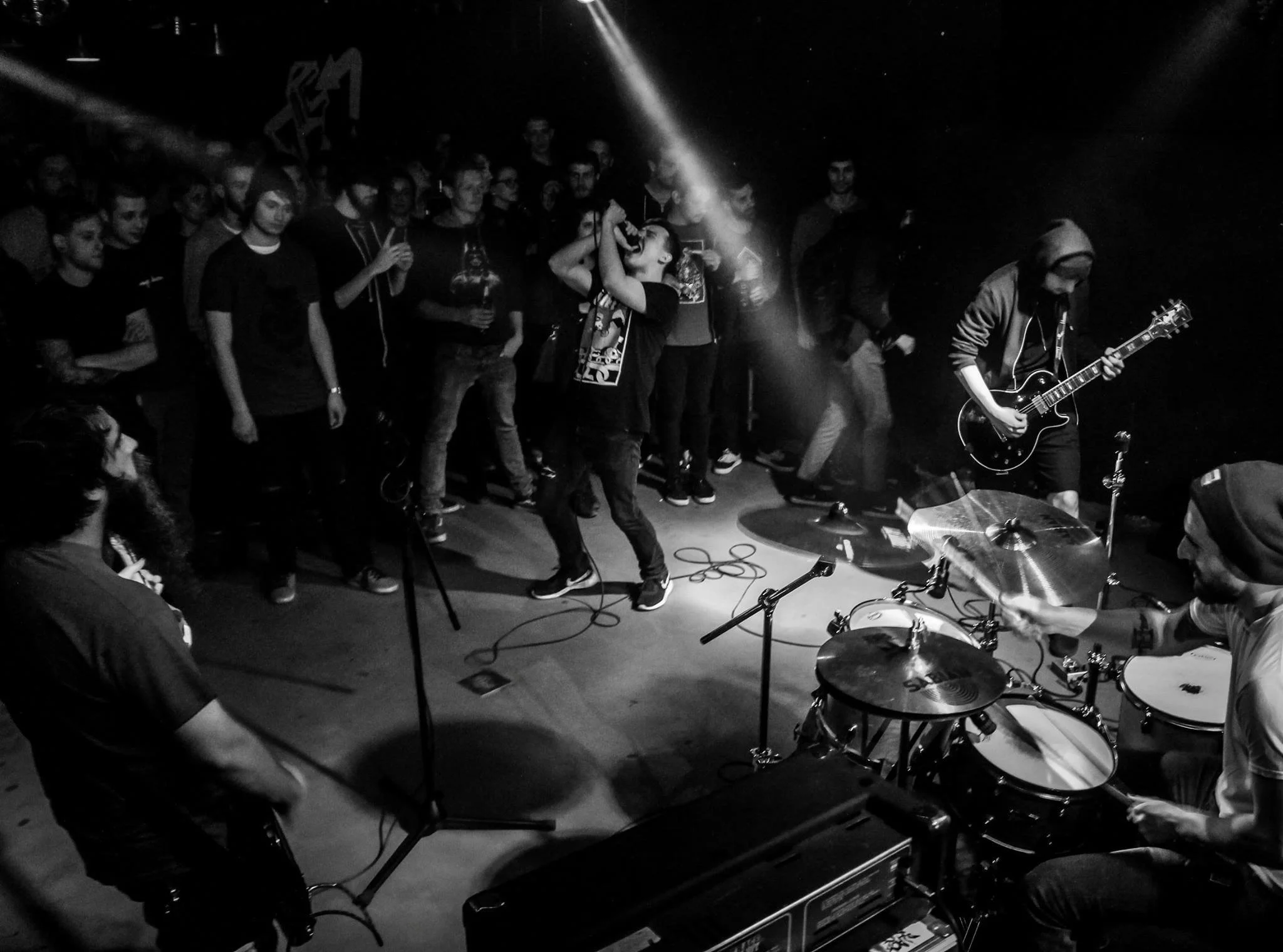 A crowded concert scene with a singer performing on stage and a guitarist playing to the right. The audience is watching intently, some with drinks, and a drummer's cymbals are visible in the foreground. The image is in black and white.