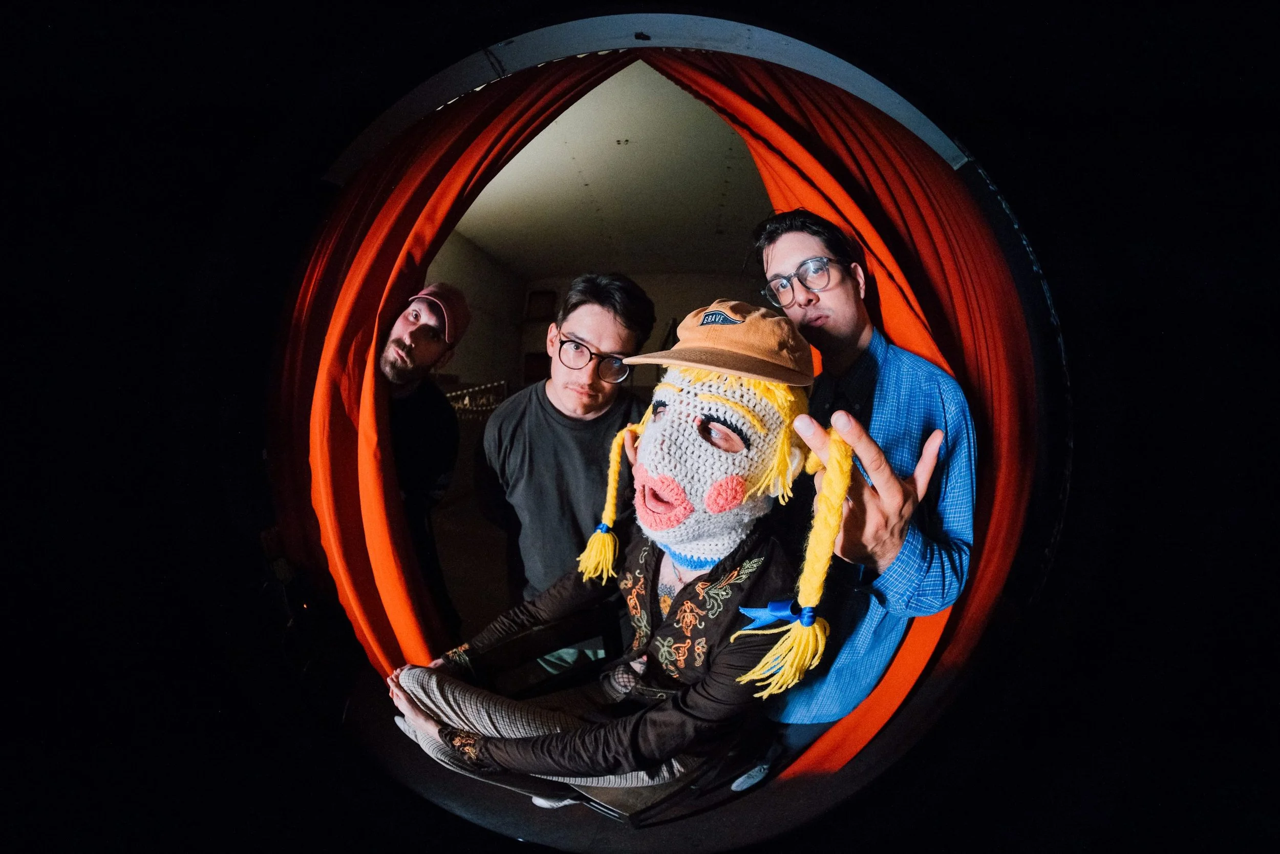 Four individuals peering into a spherical mirror, with a colorful fabric background and a person wearing a crocheted mask with a hat inside the mirror.