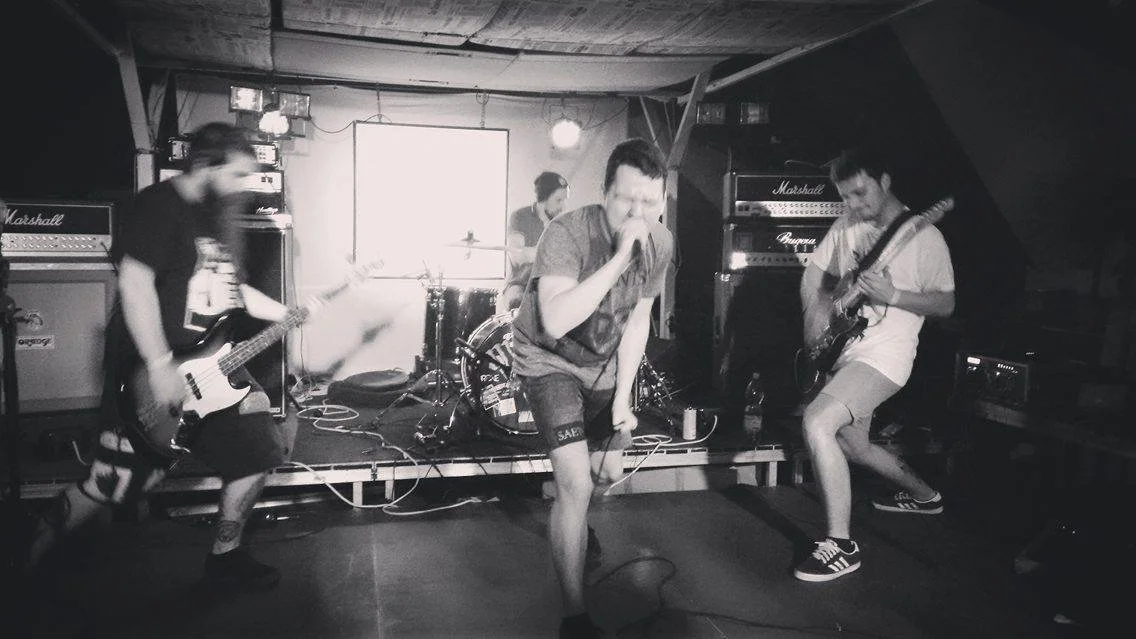 A band performing live on stage with four members playing guitars and drums, with a vocalist singing into a microphone at the center.