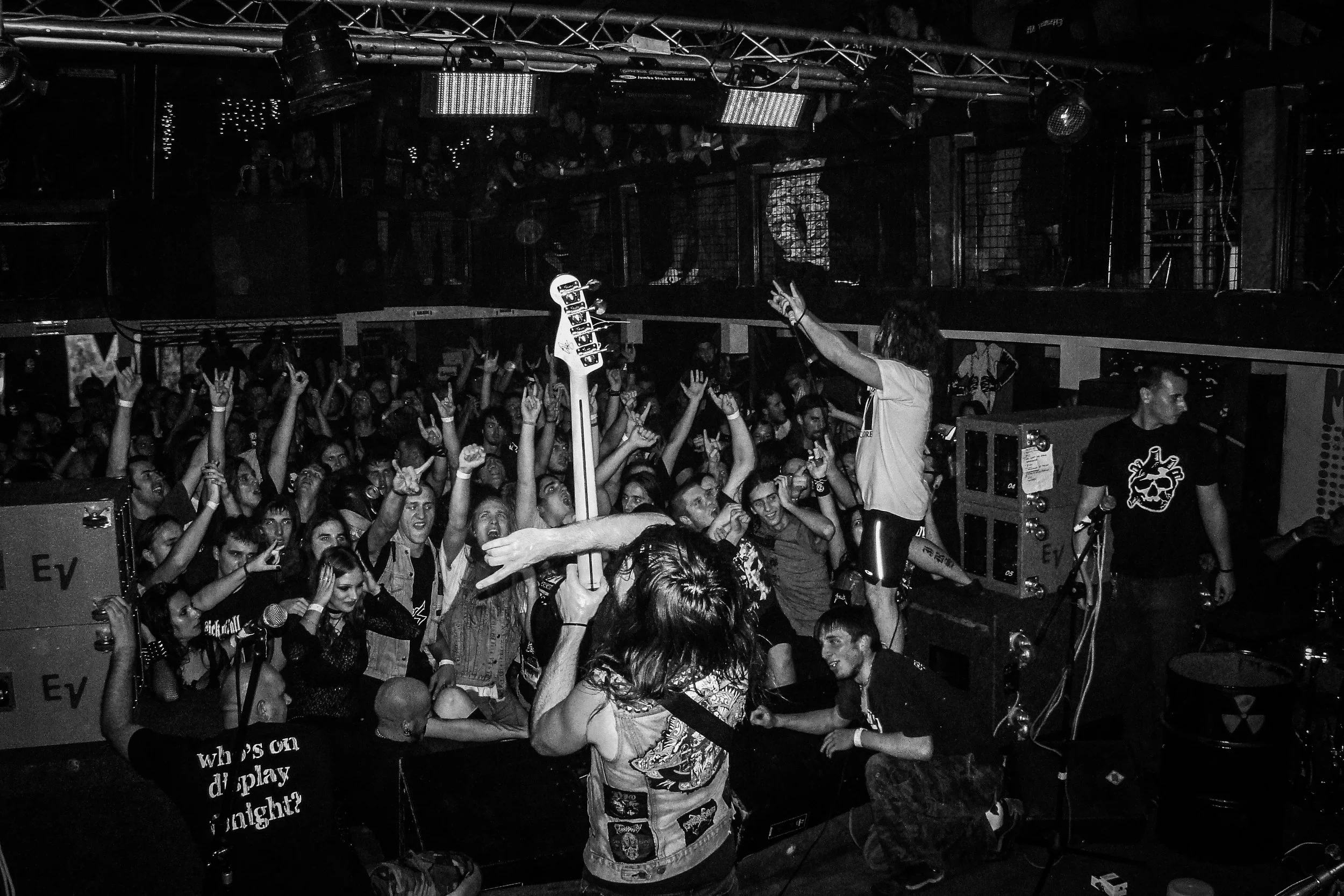 A crowded concert scene with people raising their hands, a performer with long hair singing into a microphone, and stage lighting visible overhead. Band name: Municipal Waste