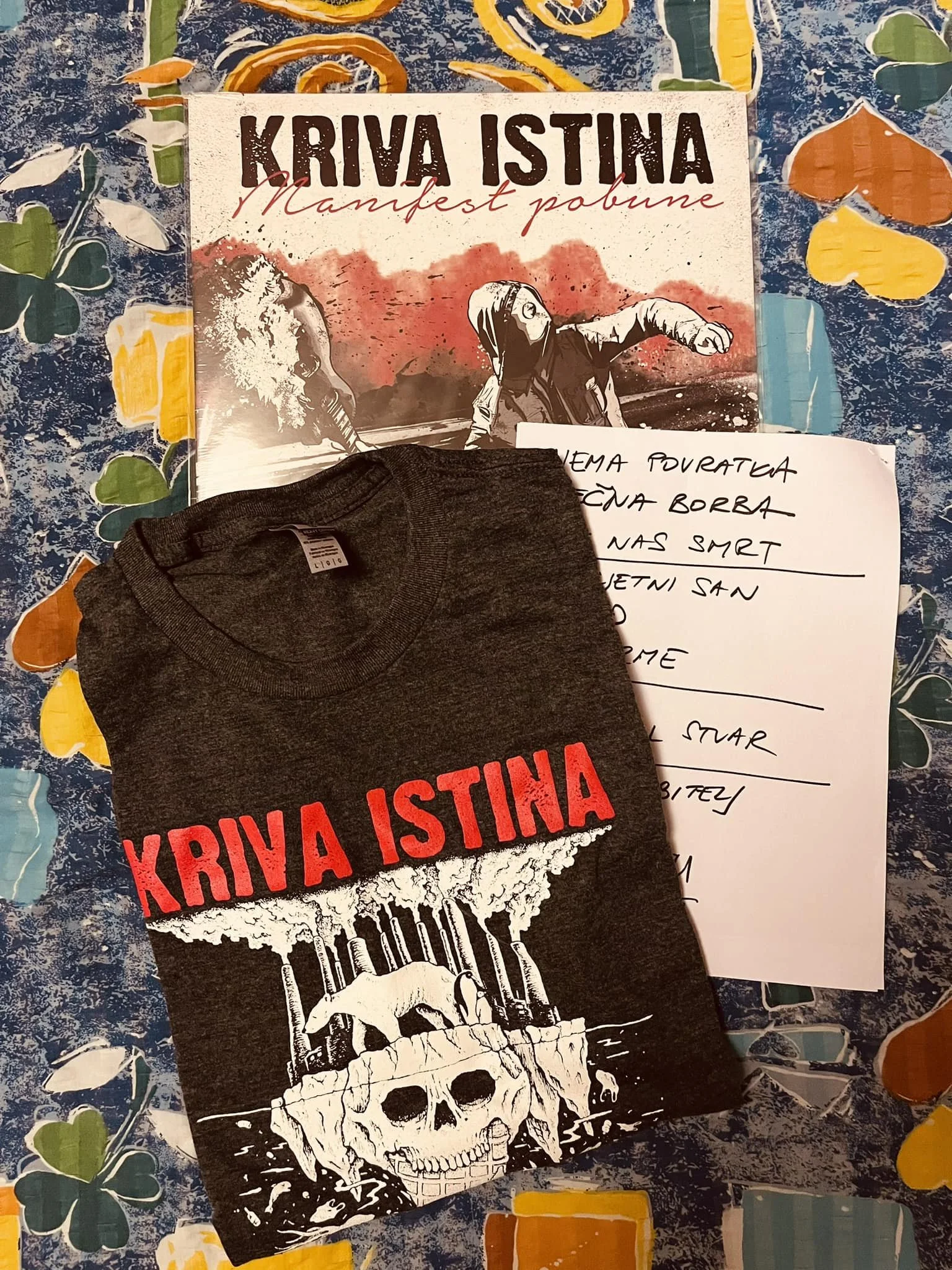 A book titled "Kriva Istina" with the subtitle "Manifest pobune" is placed on a patterned tablecloth. In front of the book, there is a folded dark gray T-shirt with red text, partially covering a piece of white paper with handwritten notes. 