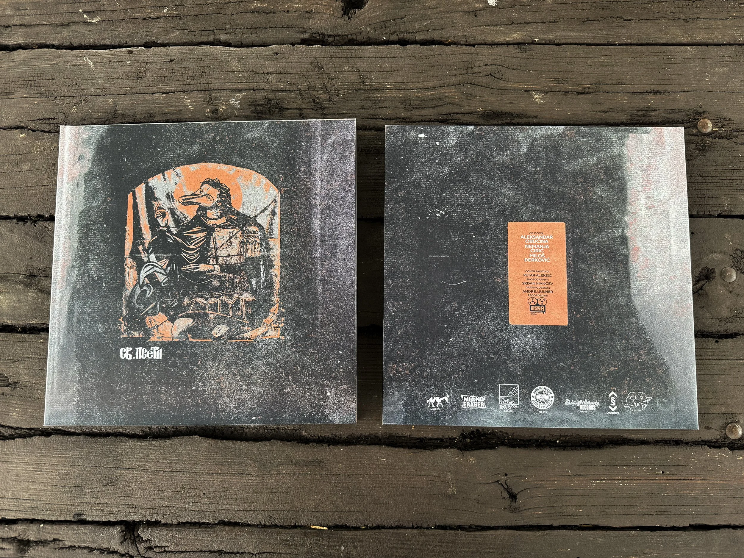 Two printed materials placed on a wooden surface. The one on the left features an illustration of a figure with a bird mask, holding a staff and standing in front of a sun or moon, with text in Cyrillic at the bottom. The second item has an orange la