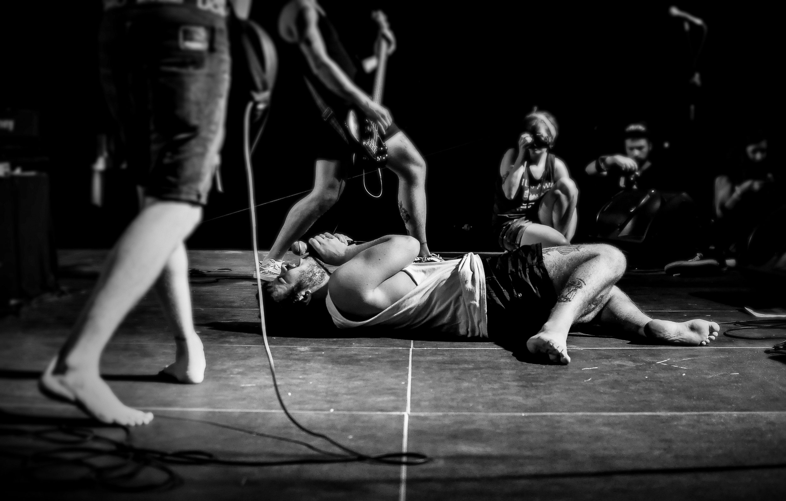 Black and white photo of a person lying on the floor with others around, possibly performing or practicing a dance or theatrical act.