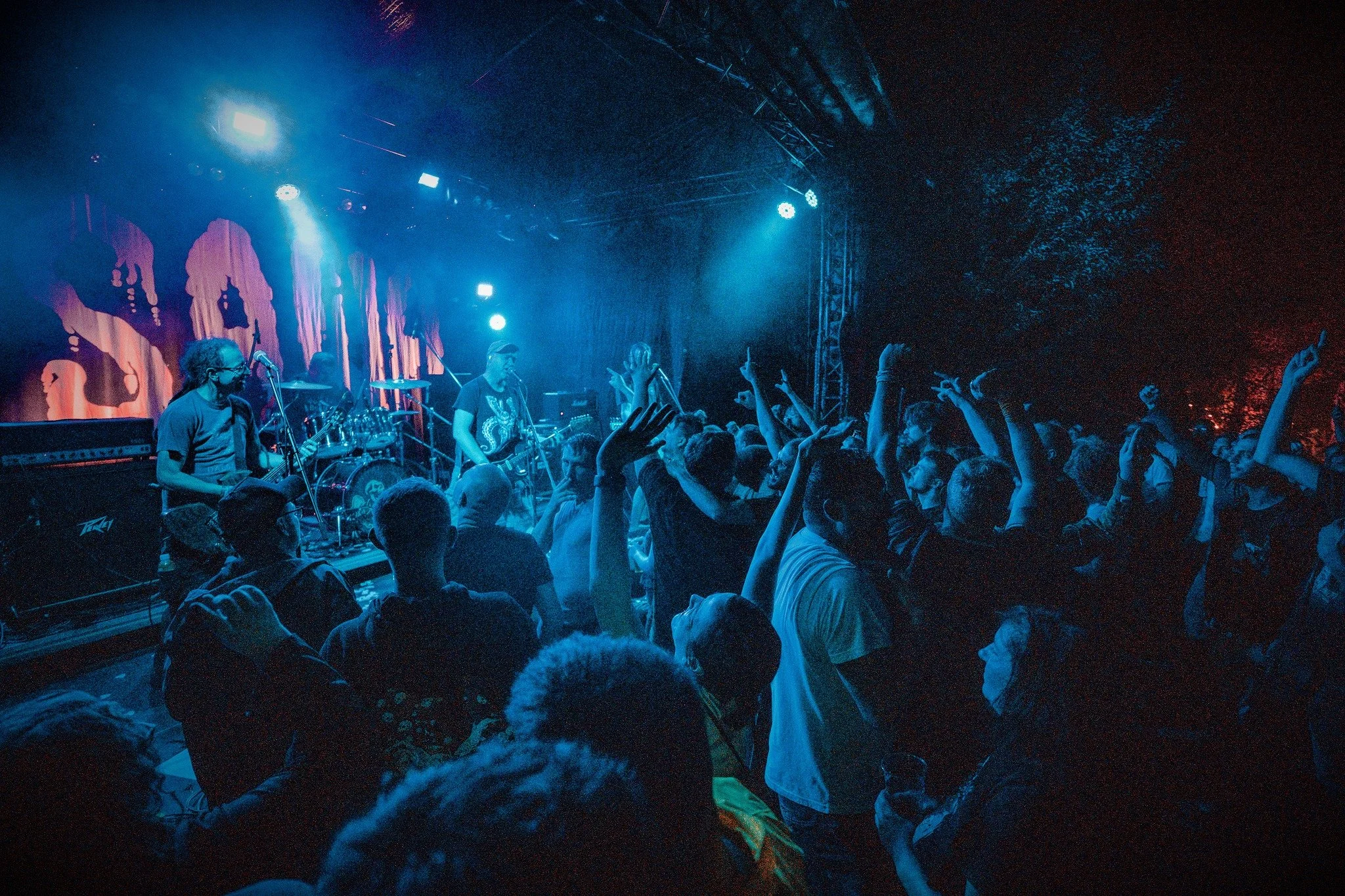 A live concert with a band performing on stage under blue lighting, audience members raising their hands, and a backdrop with silhouettes of faces.