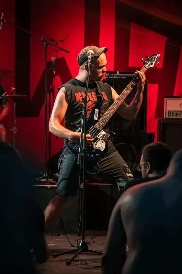 A musician playing an electric guitar on a stage with a red background, wearing a sleeveless band t-shirt and shorts, and a cap, performing for an audience.