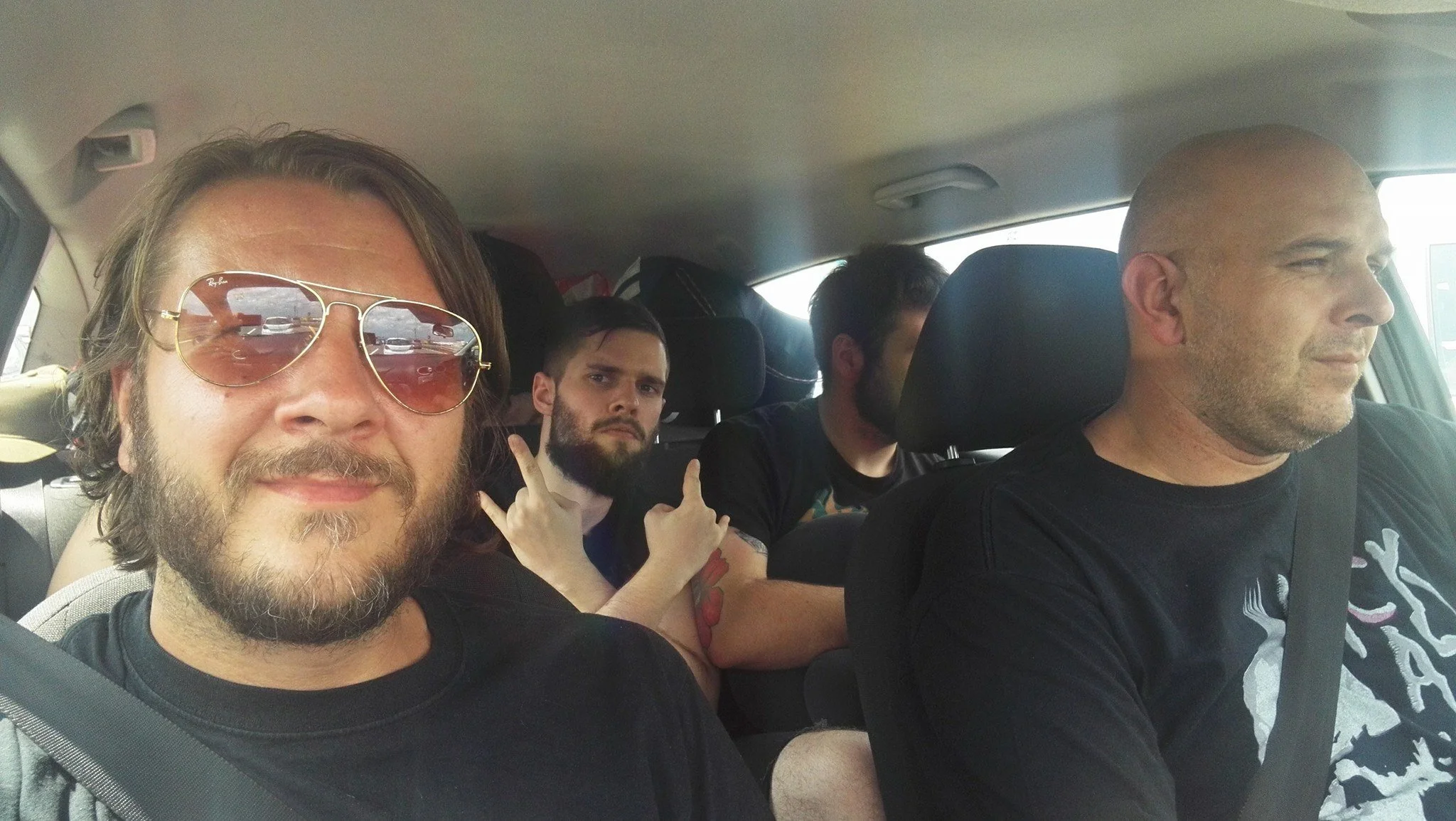 Four men sitting in a car, with the man in the front taking a selfie. The man in the front wears sunglasses, has a beard and long hair. Behind him, a young man with a beard and tattoos on his arm making a peace sign. Two other men are in the back sea