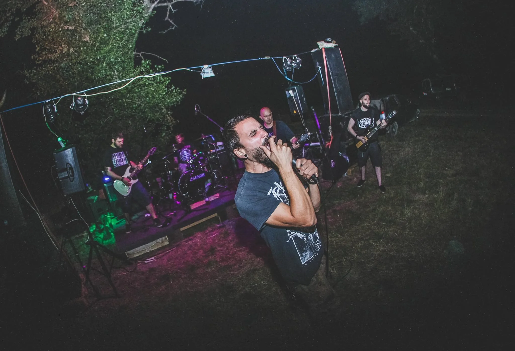 A live outdoor music concert at night with a band performing on stage. The lead singer, a man with a beard, is singing into a microphone. The band members are playing guitars and drums, and there are stage lights and equipment visible. Trees are in t