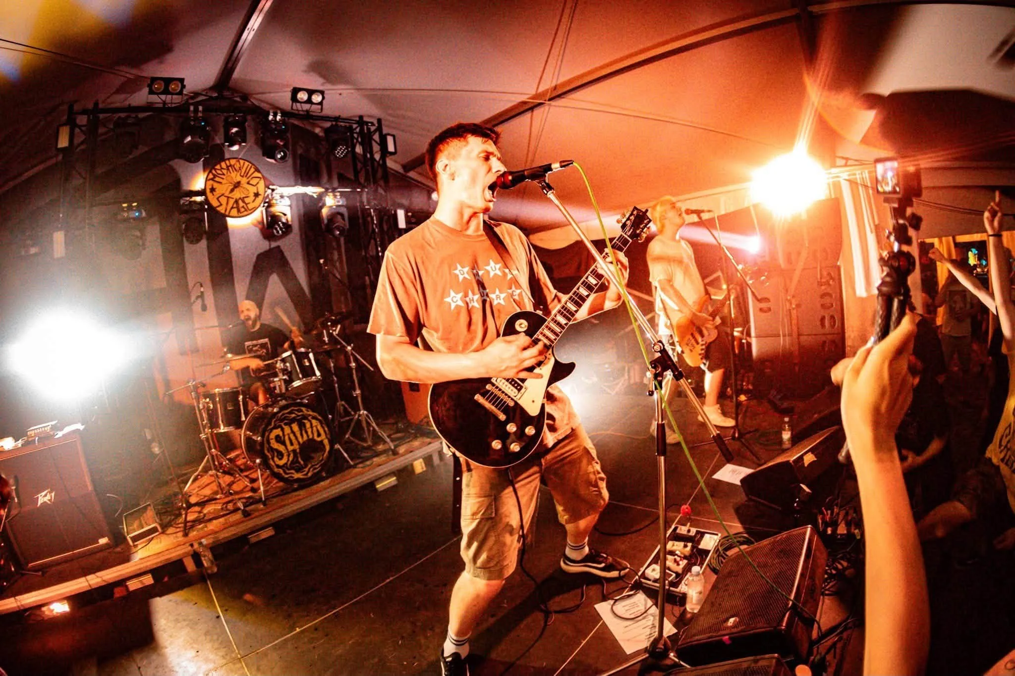 A band performs on stage at a concert, with a lead guitarist singing into a microphone, a drummer in the background, and audience members visible in the foreground.