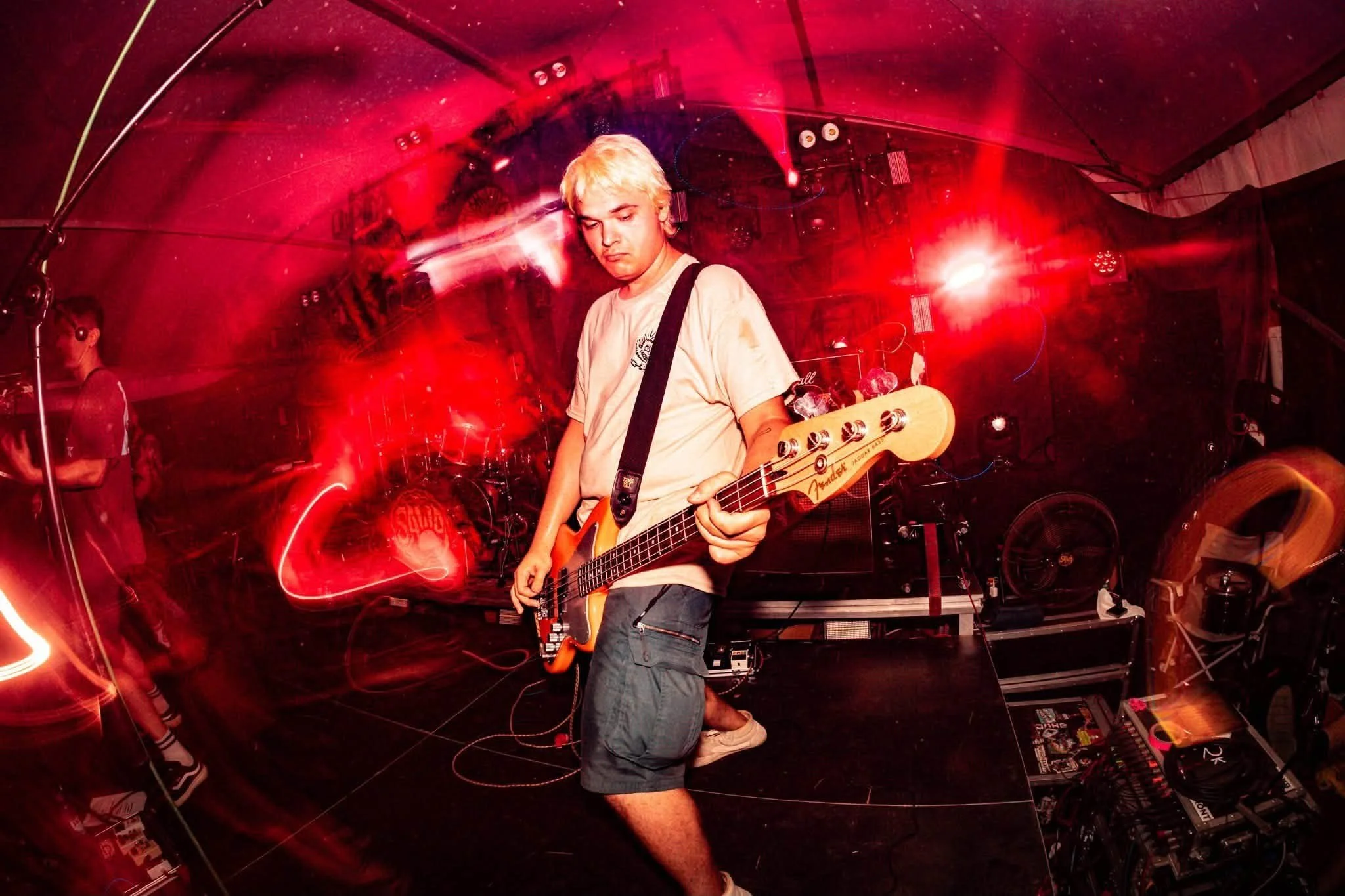 A young man with blond hair playing an electric bass guitar on stage during a live music performance, surrounded by red lighting and stage equipment.
