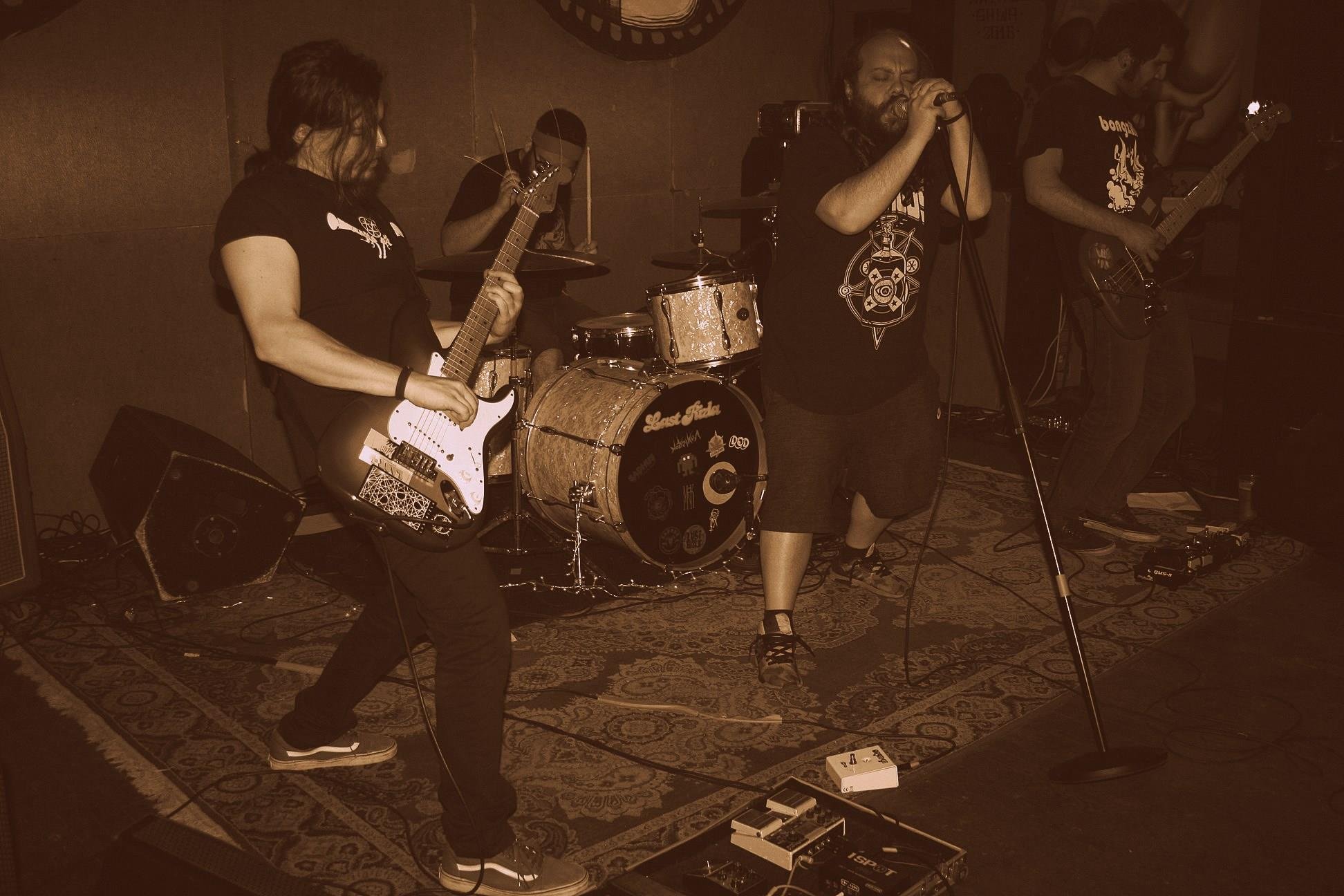 A band performing live on stage with a guitarist, drummer, female vocalist, and bassist in a dimly lit venue.