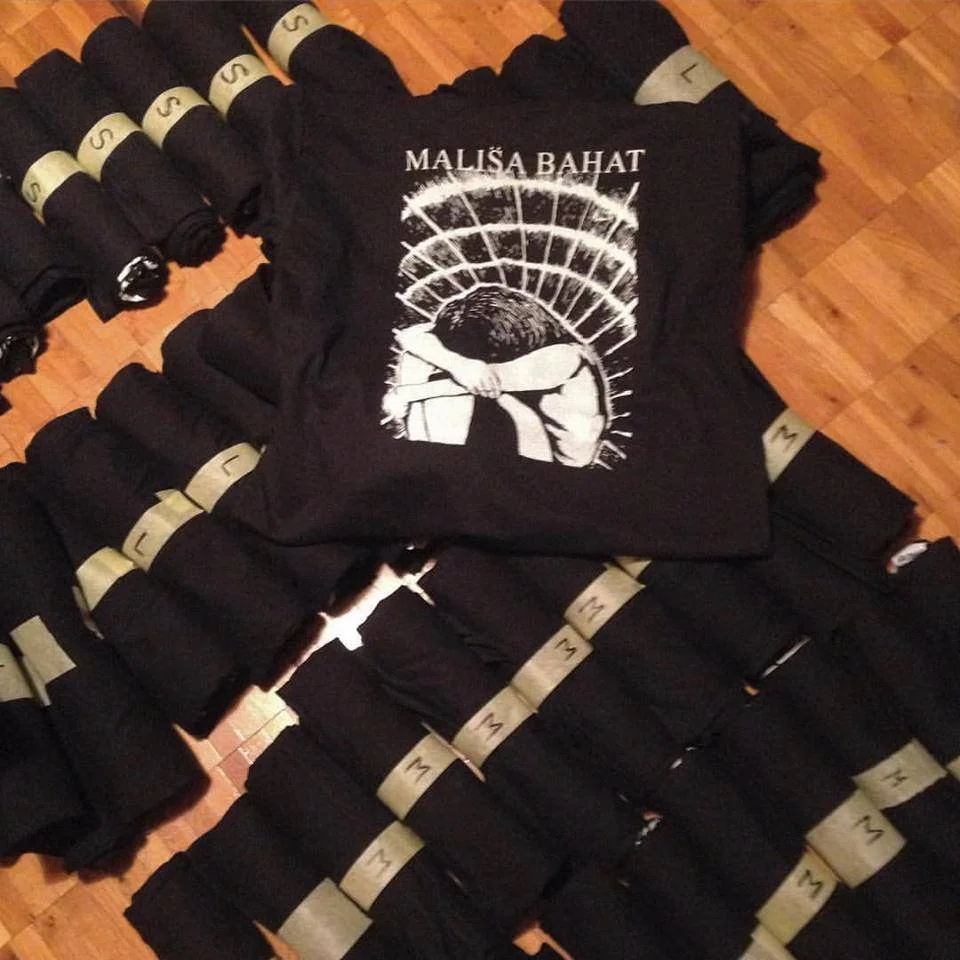 Black T-shirt with a white graphic design of a person with their head bowed, surrounded by a curved grid, and text reading "MALISA BAHAT". The T-shirt is displayed on a wooden floor among multiple rolled-up black clothing items with beige bands.