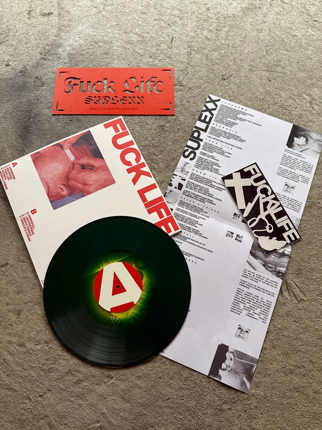 A vinyl record titled 'Fuck Life' along with a red sign, printed lyrics, and promotional material for the album 'SUPLEXX' are arranged on a textured surface.