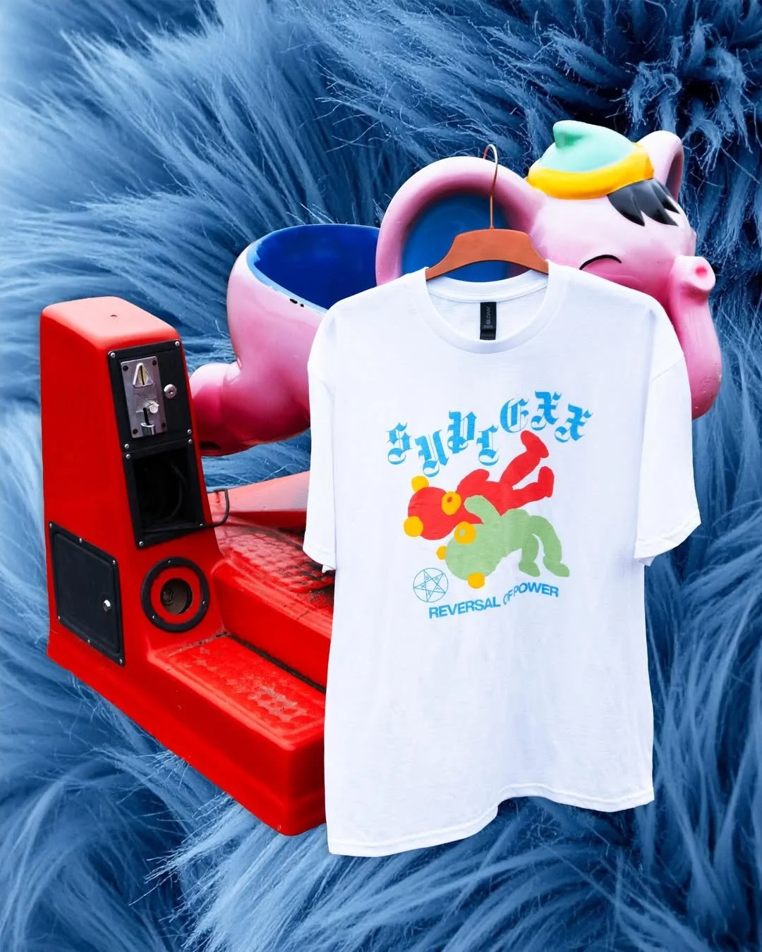 A white T-shirt with colorful graphic design and text hangs on a hanger. The background features a large pink and blue unicorn sculpture and a red vintage-style radio or speaker.