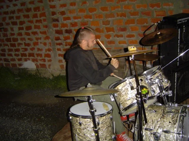 A man with a mohawk hairstyle playing a drum set outdoors at night, with a brick wall in the background.