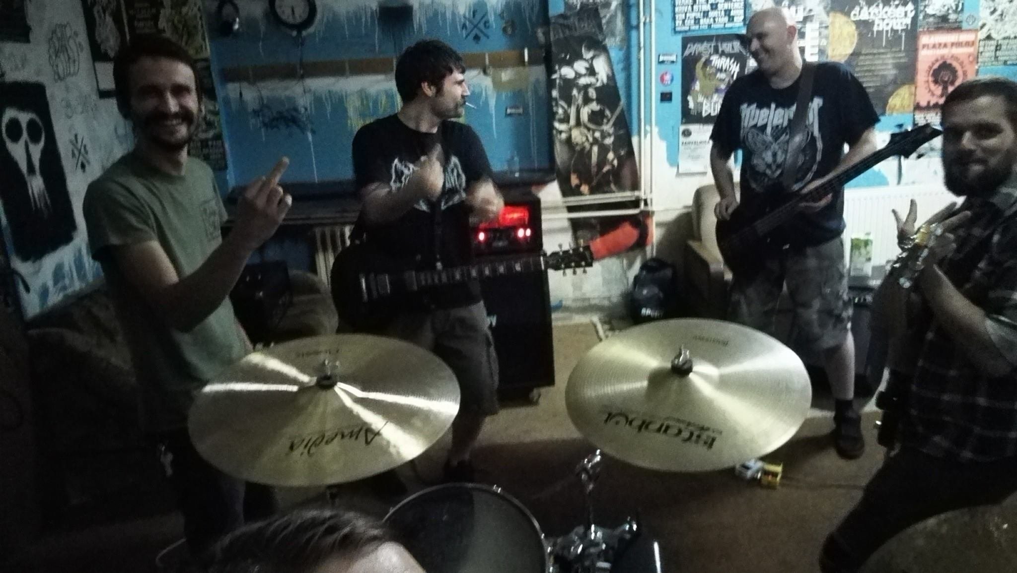 Four men playing musical instruments in a dimly lit room with graffiti-covered walls. One holding a guitar, one with a microphone, one with a bass guitar, and another with drums.