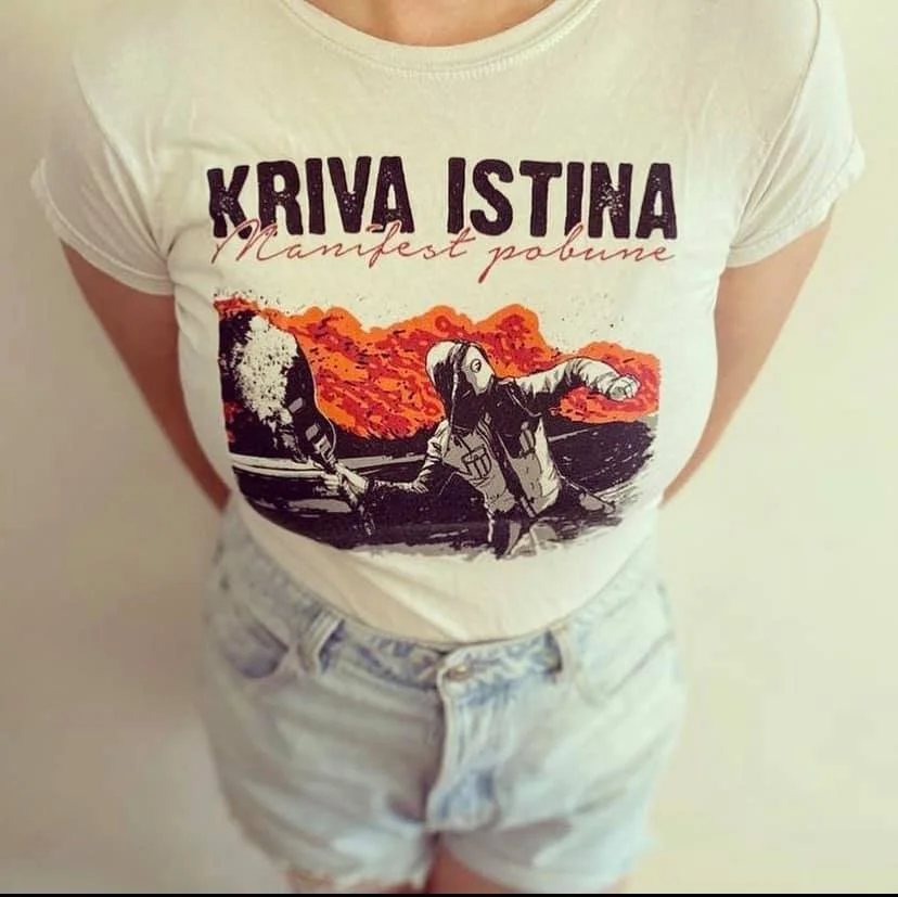 A person wearing a white T-shirt with a graphic of a woman in a vest pointing forward, with a background of orange and black flames, and text that reads 'KRIVA ISTINA' and 'Manifest pobune'. The person is standing with arms behind their back, wearing