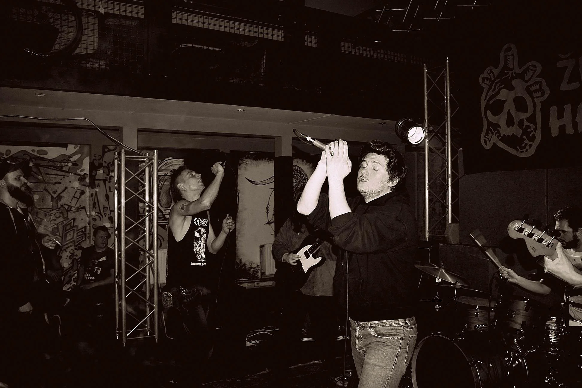 A group of musicians and singers performing on stage in a music venue. Some are playing guitars, others are singing, and one person in front is holding a microphone with his eyes closed.