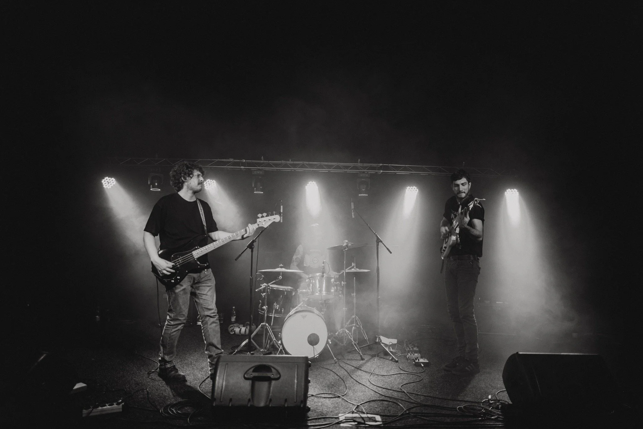 A black-and-white photo of a band performing on stage, with two guitarists playing their instruments and a drummer in the background. The stage is lit with bright lights from above.