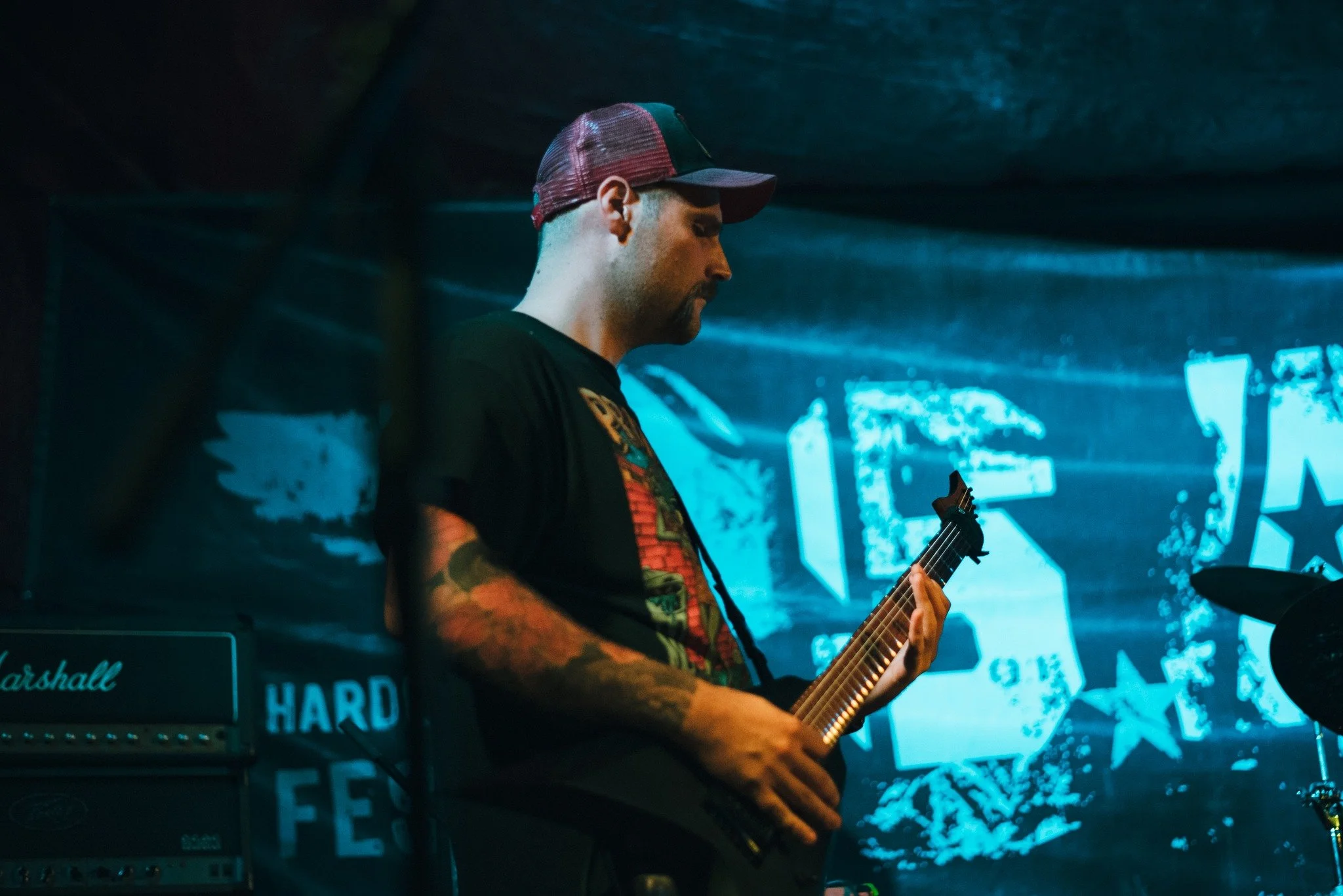 A man with tattoos on his arms wearing a baseball cap and black T-shirt playing an electric guitar on stage, with a large screen in the background displaying graphics and text.