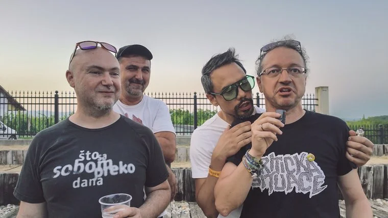 Four band member from LOS PRIMJER standing outdoors near a black fence. They are casually dressed, with two wearing glasses. One man in front is holding a plastic cup, and another is leaning on him while holding a small device.
