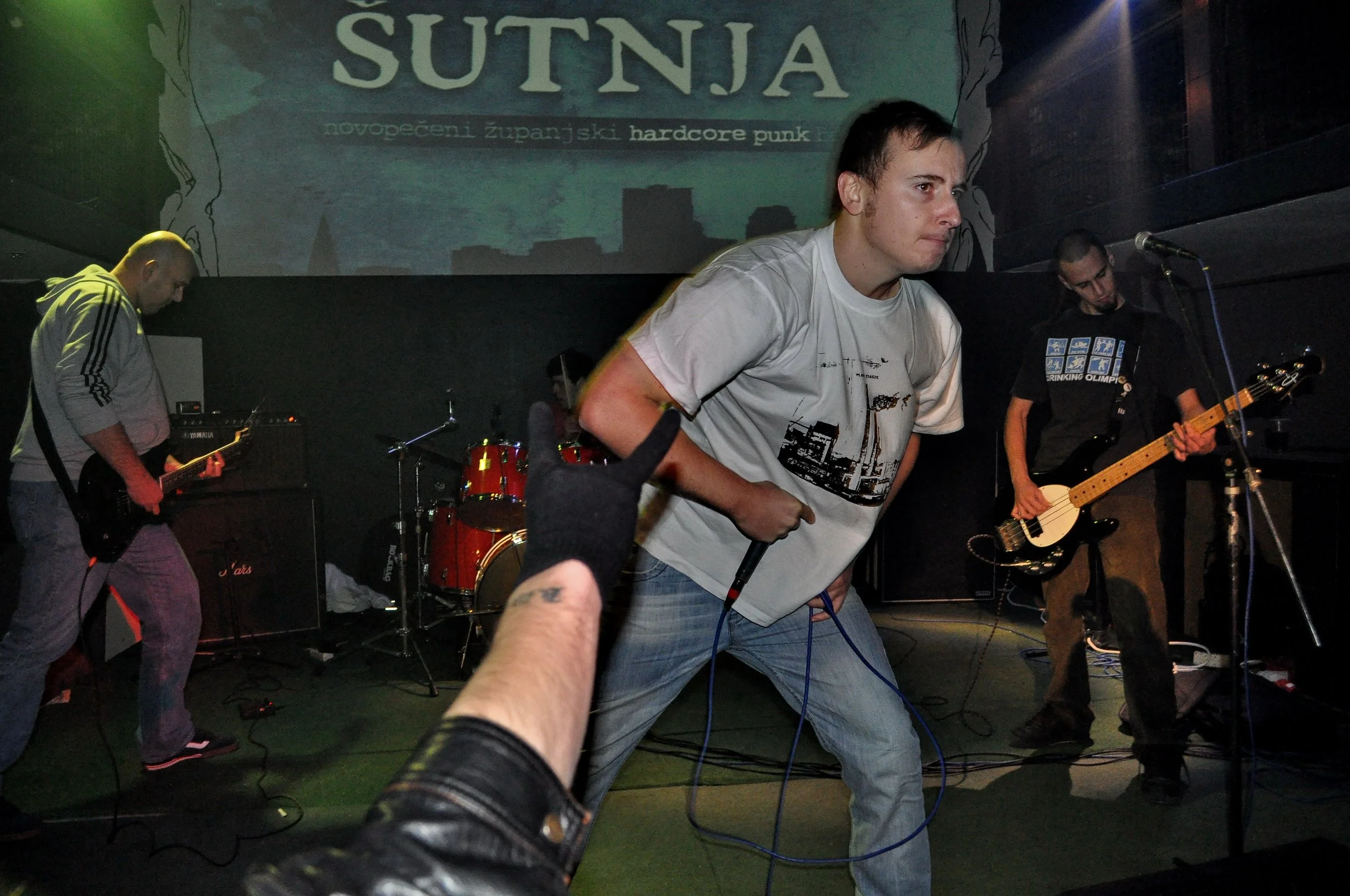 Band performing on stage with a singer in front, backup musicians playing guitars, drums and bass, and a large screen with text in the background.