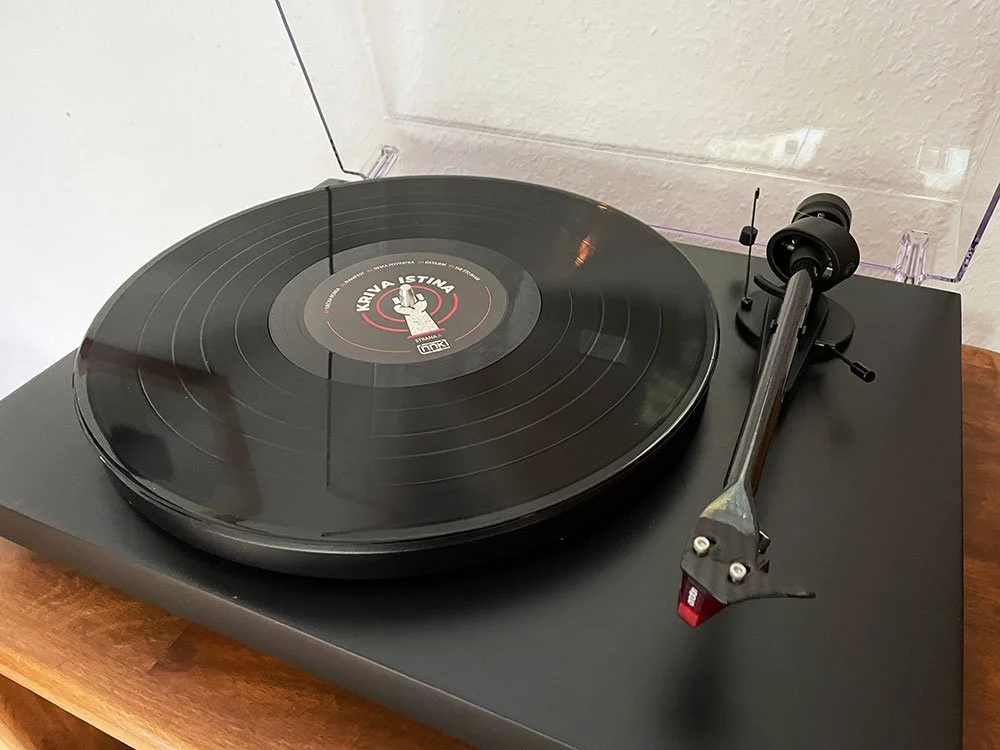 Black vintage turntable with a vinyl record on it, placed on a wooden table in a room with white walls.