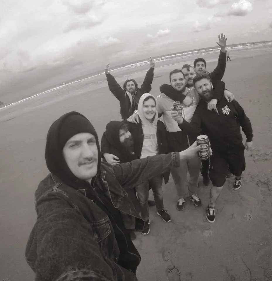 Group of eight friends taking a selfie on a beach, with some holding drinks, under cloudy skies.