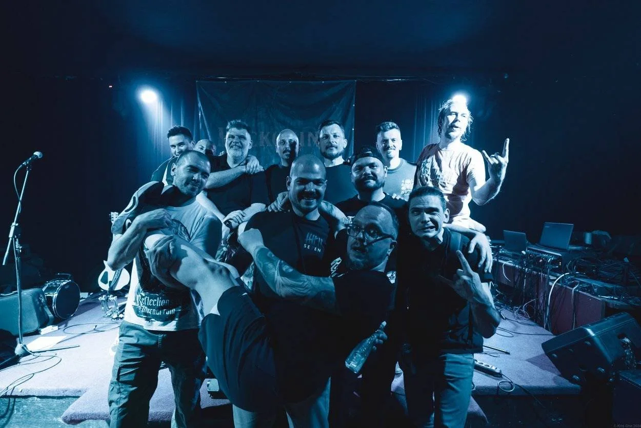 Group of people on stage at a concert or event, some smiling and showing hand gestures, with musical equipment and a banner in the background.