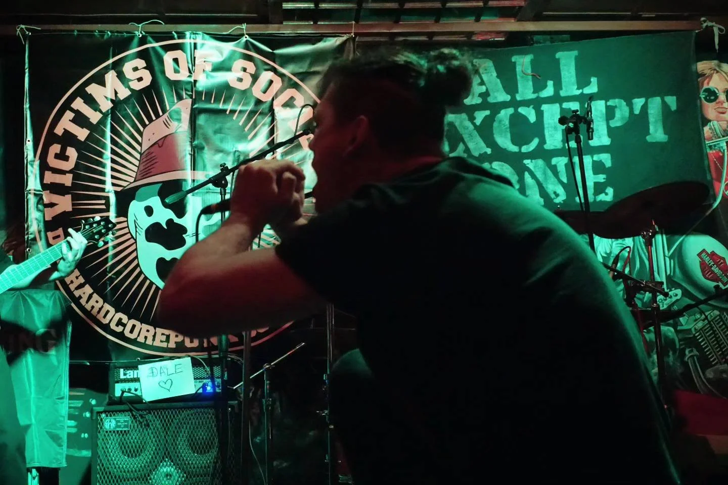 A band performing on stage at a lively nightclub, with drummer, guitarist, and singer under colorful stage lighting and a backdrop with slogans about hardcore punk and DC.
