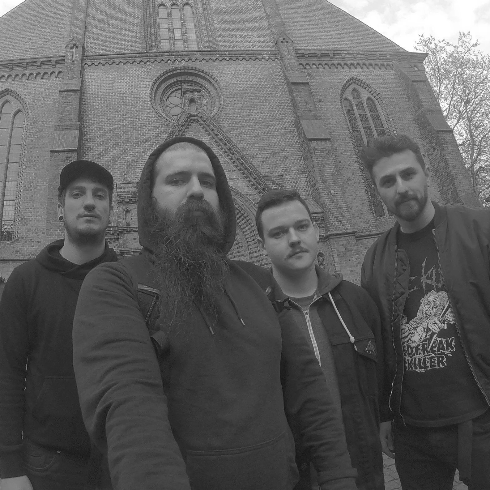 Four men taking a selfie in front of a large brick church with stained glass windows, trees, and cloudy sky in the background.