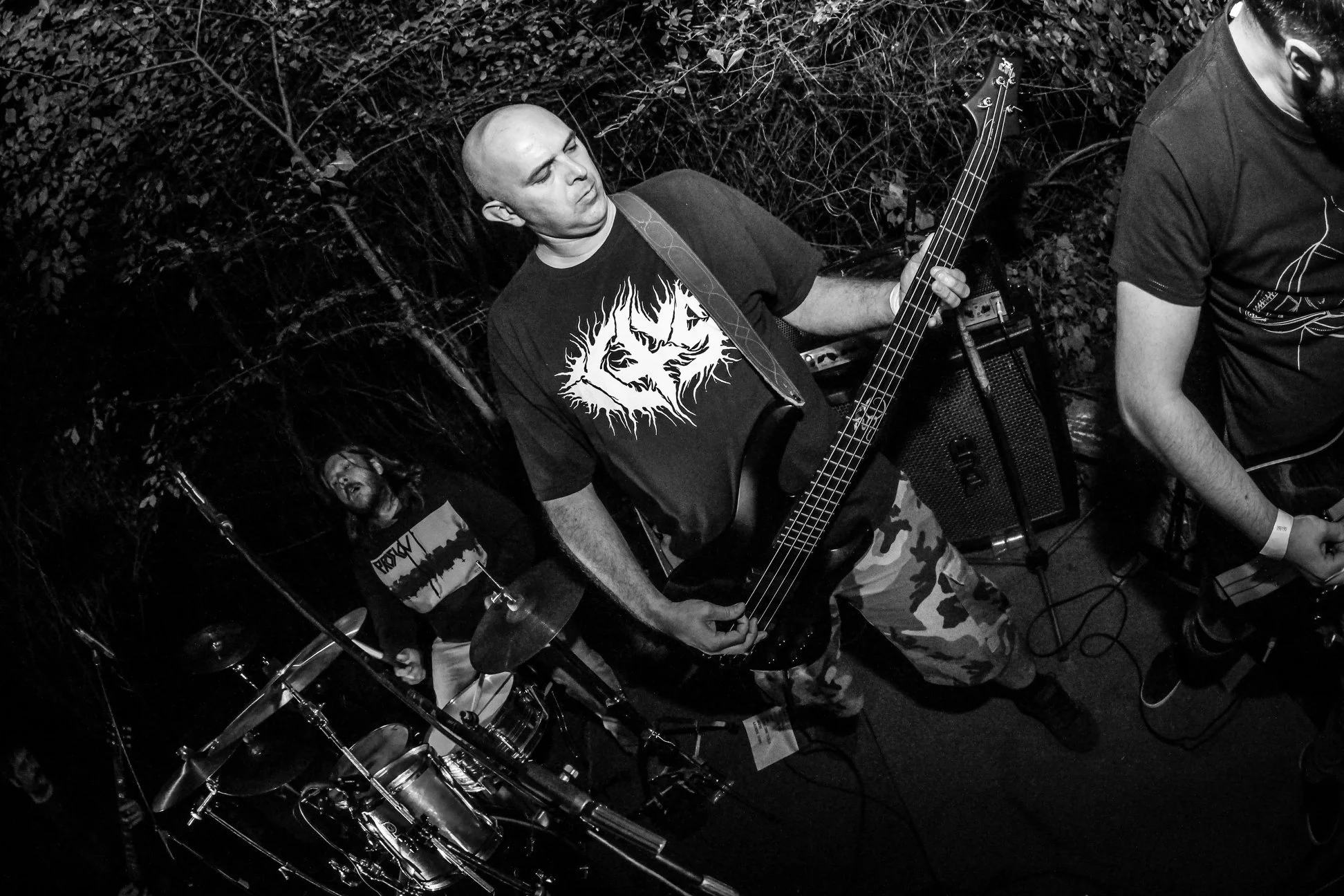 Black and white photo of a music band performing outdoors. A bald man is playing an electric bass guitar, wearing a dark t-shirt with a graphic design. There is a drummer in the background with a graphic t-shirt. The scene is set amid dense foliage.