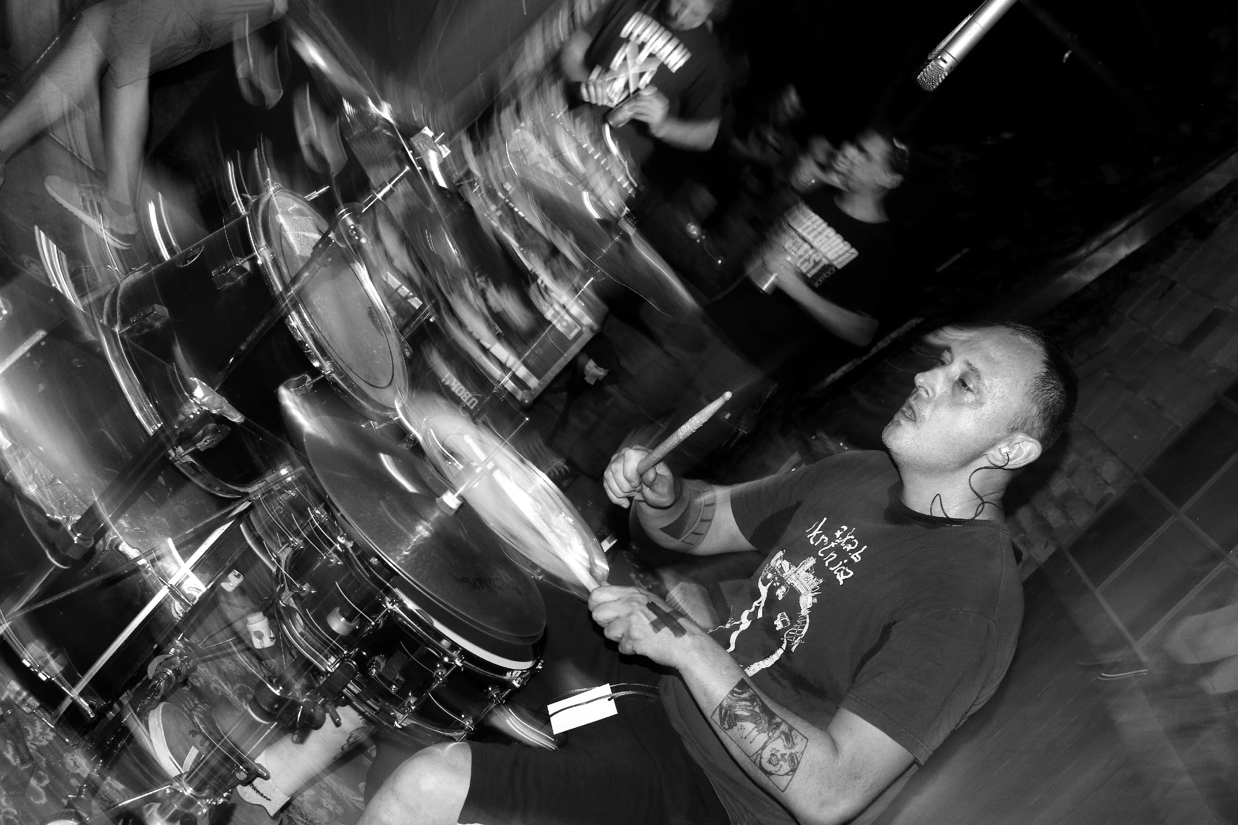 A person with tattoos on their arm and earrings is playing the drums in a dimly lit room. There are other people and music equipment visible in the background.