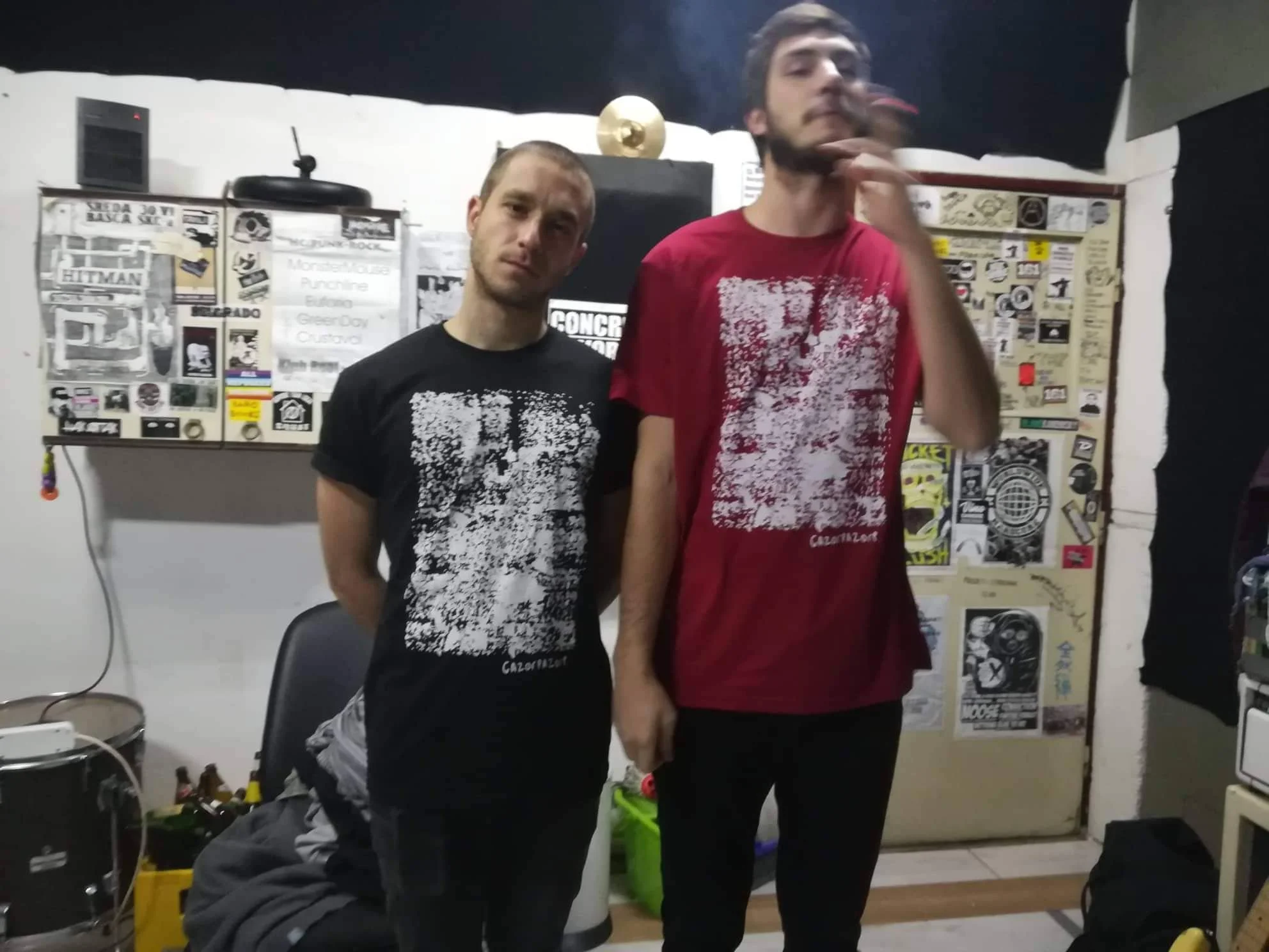 Two young men standing side by side in a room with white walls and decorated with various posters and stickers. One is wearing a black t-shirt, and the other is wearing a red t-shirt. They are near a black chair and a white appliance, possibly a refr