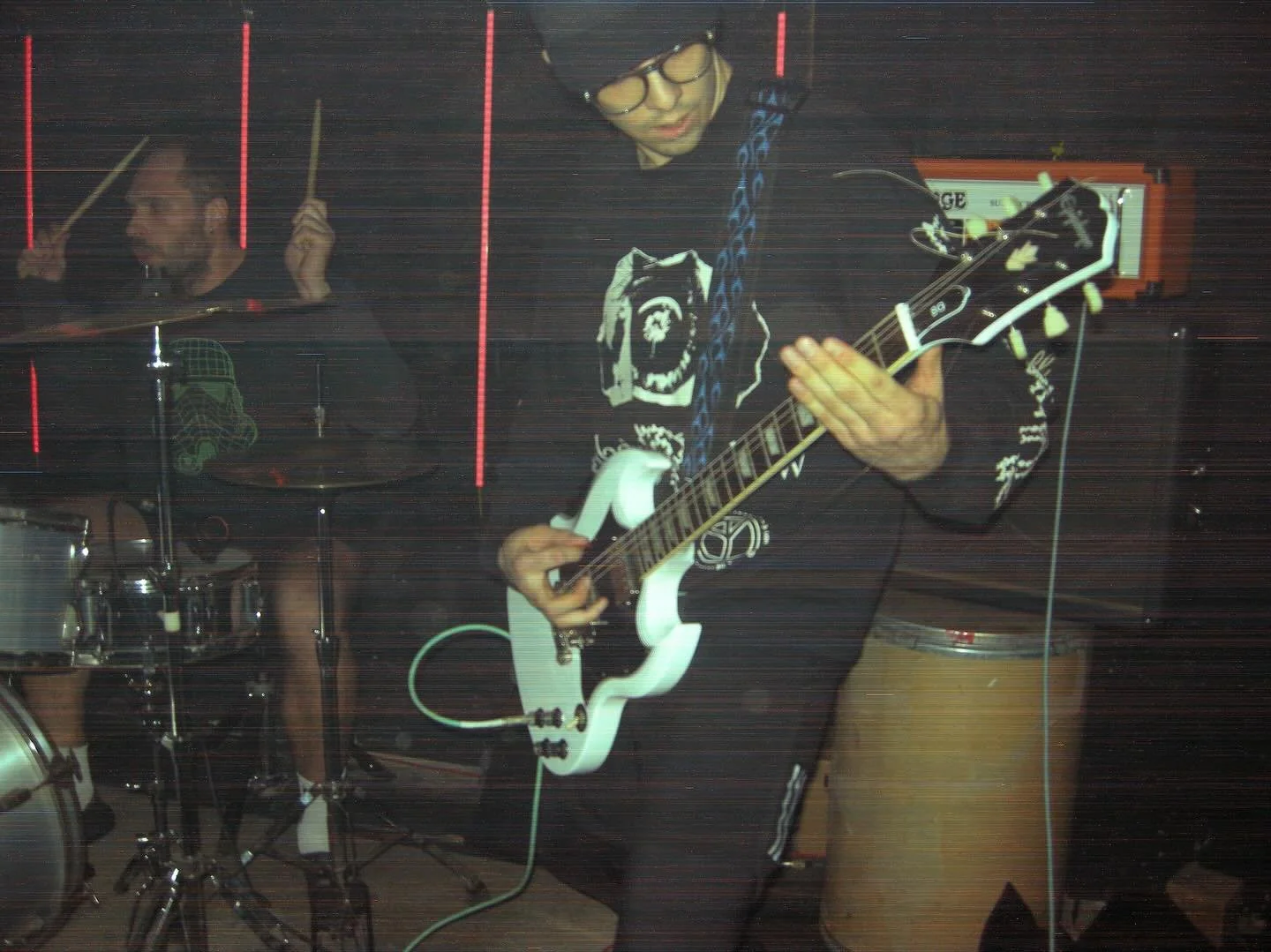 Musicians playing instruments, a drummer and a guitarist, in a dimly lit room with red vertical light lines.