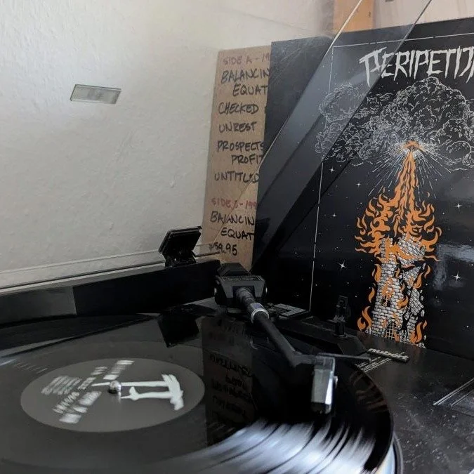 A vinyl record player is playing a spinning black vinyl record. In the background, there is a room with a white textured ceiling and a brown cardboard sign with handwritten notes. To the right, there is a black turntable cover or poster with a fiery 