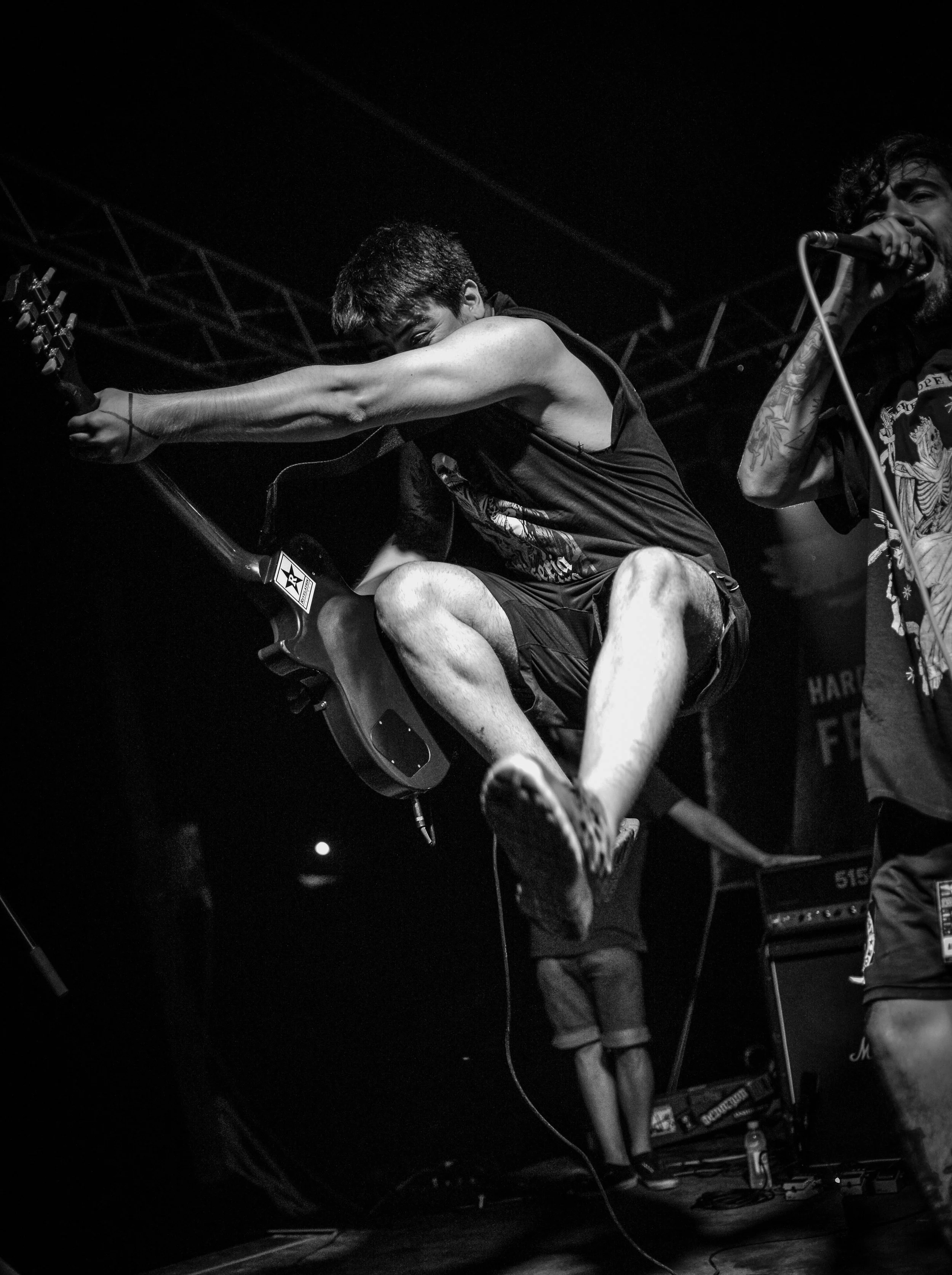 Black and white photo of a young man jumping on stage playing an electric guitar with one arm stretched out. Another musician on the right, singing into a microphone. Musical equipment is seen in the background. Band name: En Mi Defensa