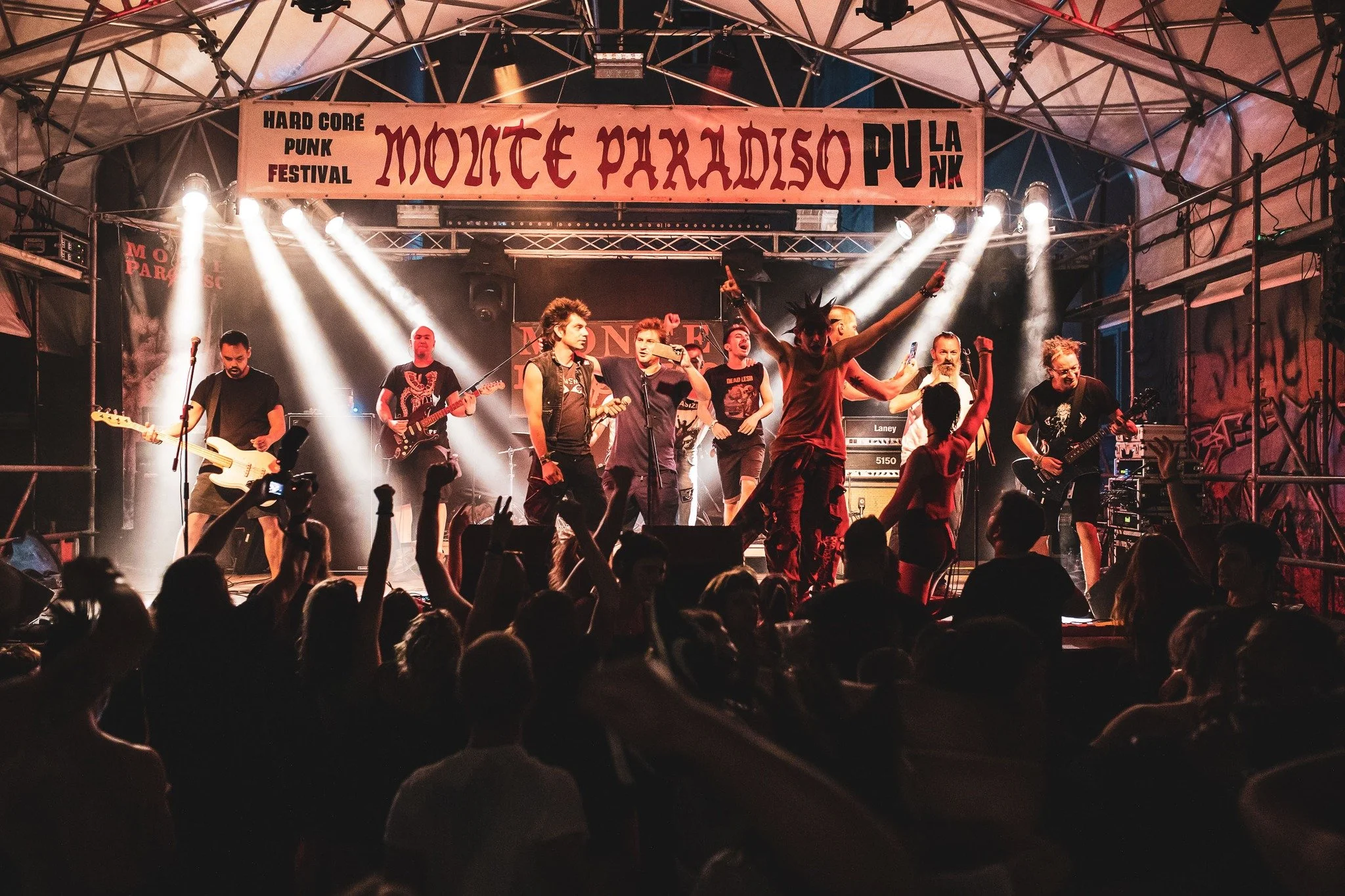 A live outdoor punk music performance at the Monte Paradiso Punk Festival, with a band on stage, audience members raising their hands, and stage lighting illuminating the performers under a large banner that reads 'Monte Paradiso Punk Festival'.