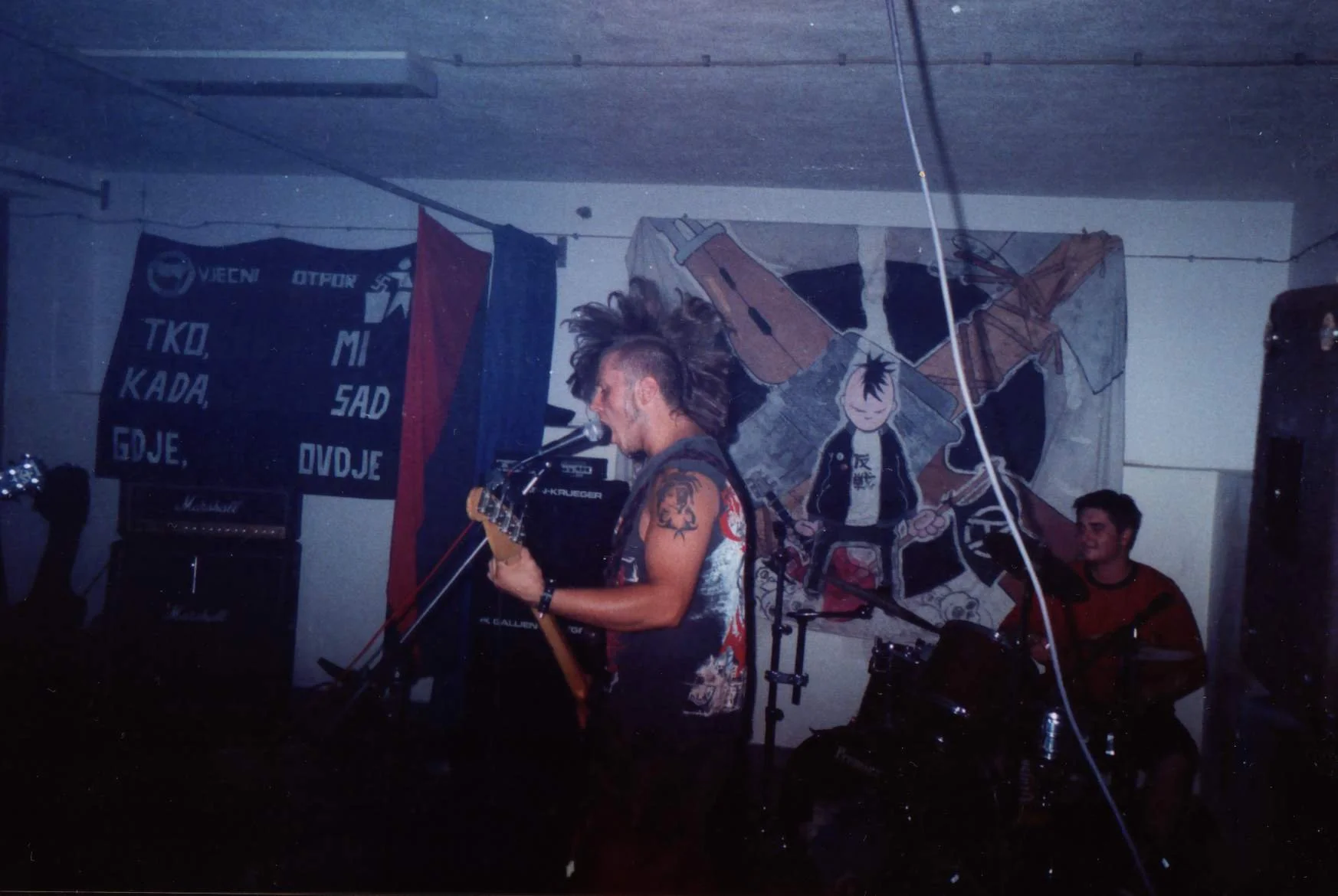 A musician with a mohawk playing a guitar and singing into a microphone at a small indoor concert. A drummer is seen in the background, and the walls are decorated with flags and artwork.