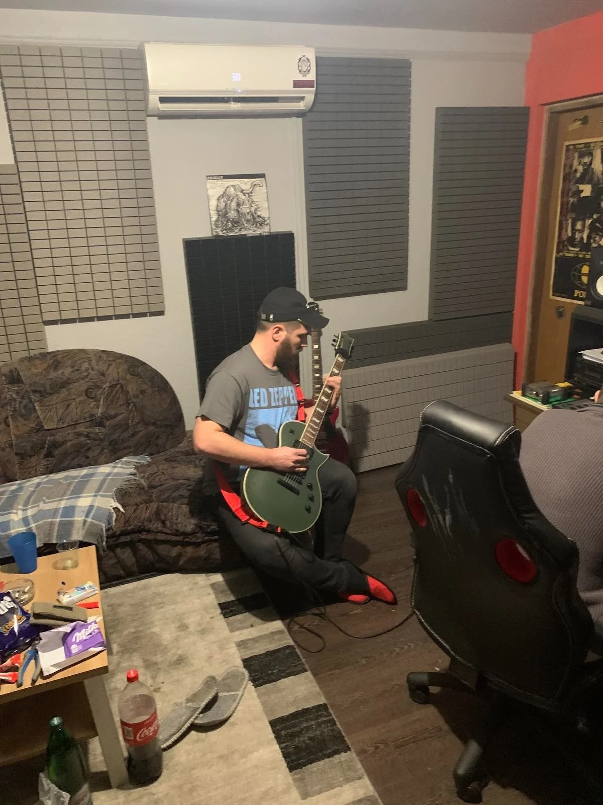 A man with a beard and black cap playing an electric guitar in a music studio or room. The room has soundproof foam panels on the wall, a brown patterned couch, a small table with snacks and drinks, and various equipment and posters.