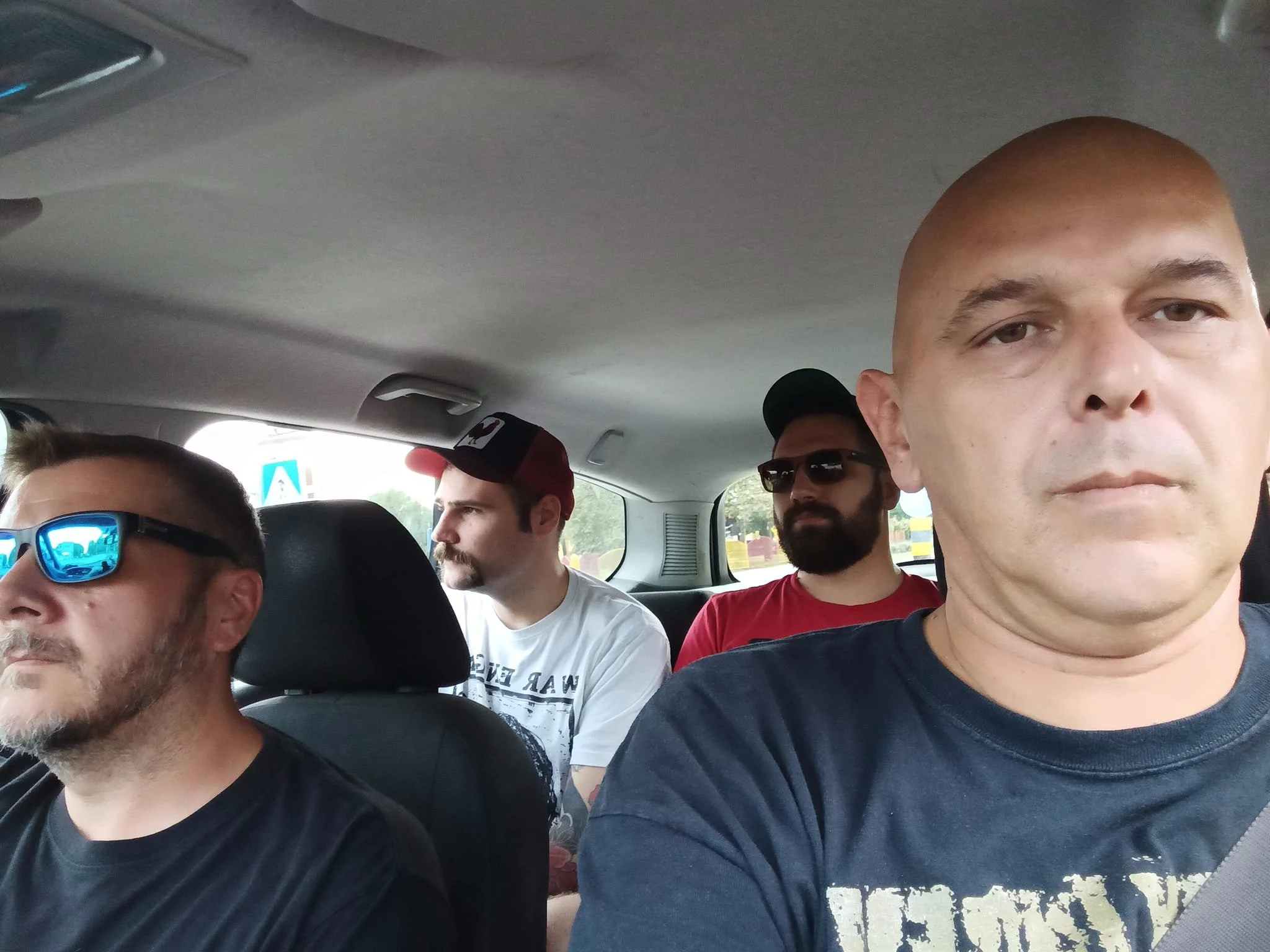 Four men sitting in the backseat of a car with one man in the front taking a selfie. 