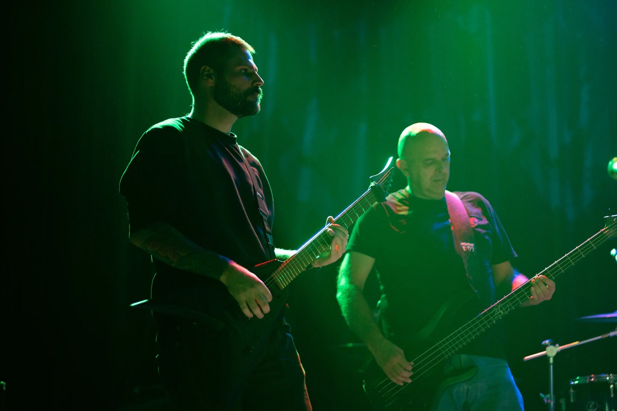 Two musicians are playing electric guitars on stage, illuminated by green lighting. One is a bearded man with a tattooed arm, and the other is a bald man. Both are focused on their music.
