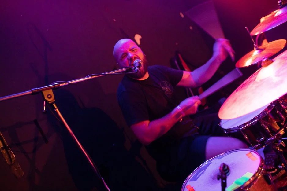 A male drummer passionately playing drums and singing into a microphone during a live performance in a dimly lit venue.