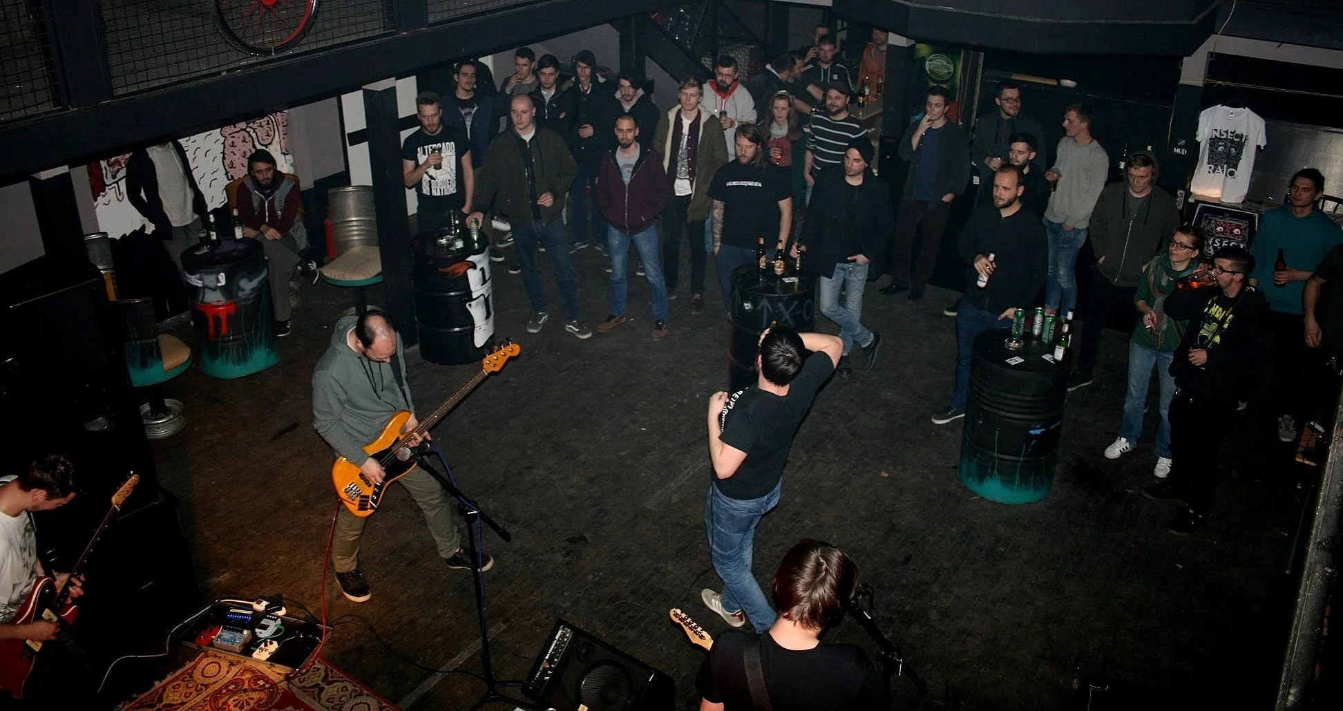A live music performance at a small venue with a band playing guitars and a bassist. The audience is standing and watching, some holding drinks. The venue has a dark, industrial style with barrels and graffiti on the walls.