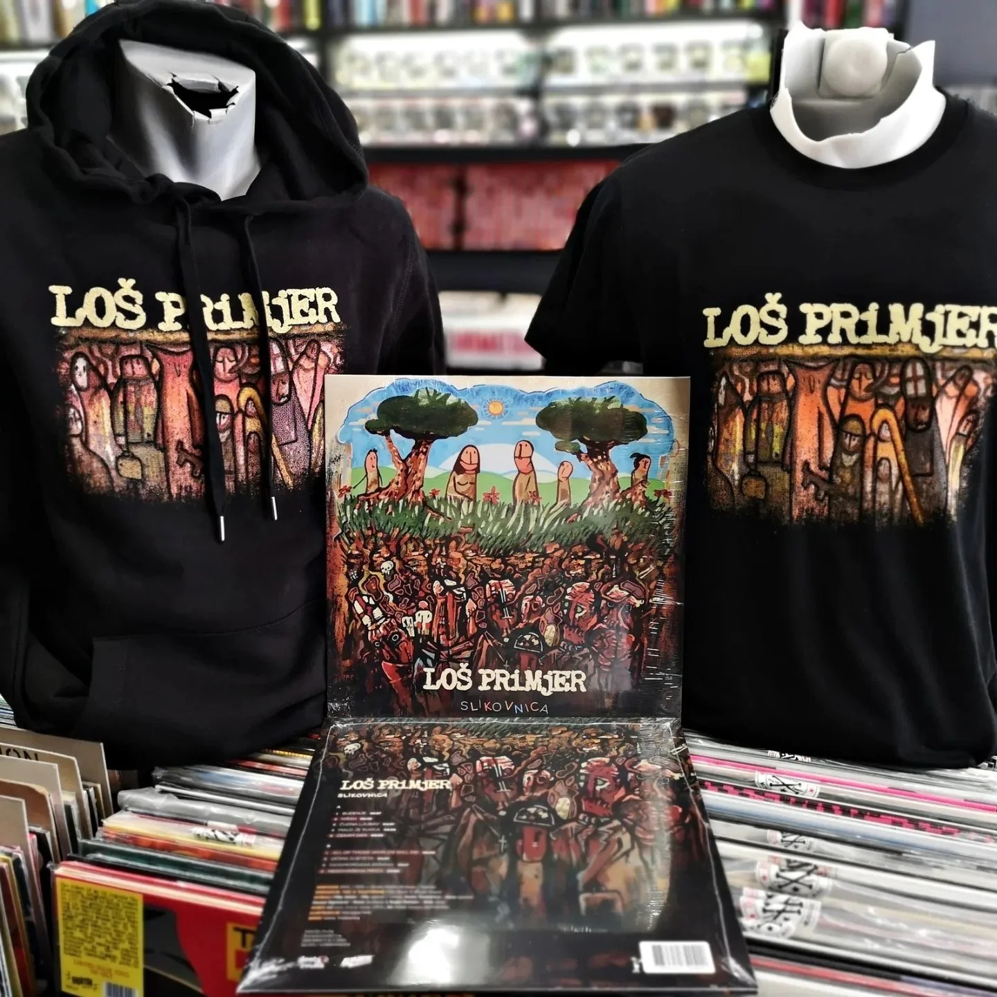 A display of black hoodies and t-shirts with a colorful graphic design and text that reads "LOS PRIMJER" in a store. The graphic depicts stylized trees, human figures, and abstract patterns, with books and magazines underneath.