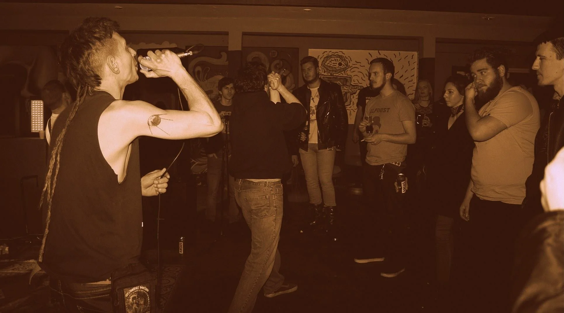 A person with dreadlocks singing into a microphone at a crowded indoor concert or event, with audience members standing and watching.