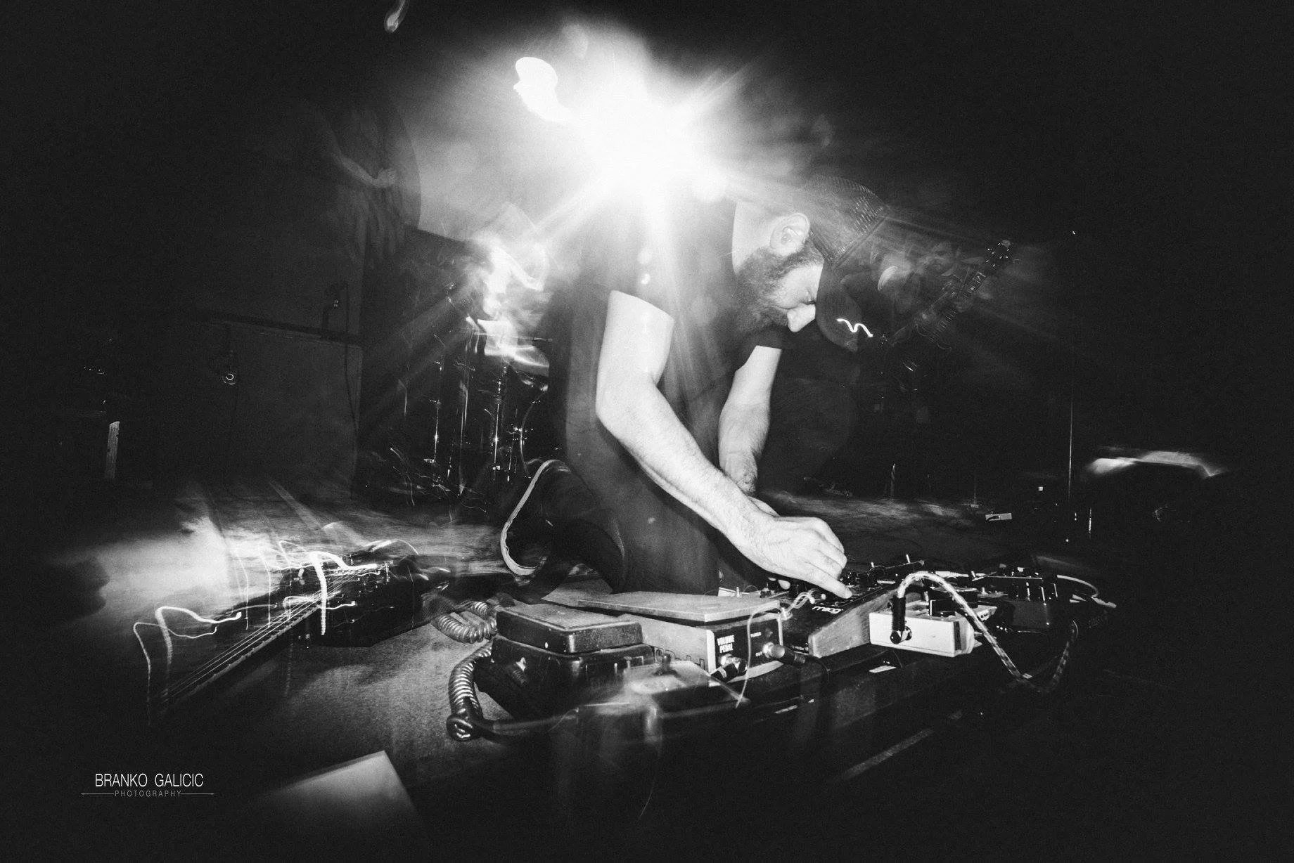 A male DJ with a beard, wearing a cap, is performing at a club or music event, kneeling and manipulating DJ equipment, with intense lighting and motion effects creating a dynamic atmosphere.