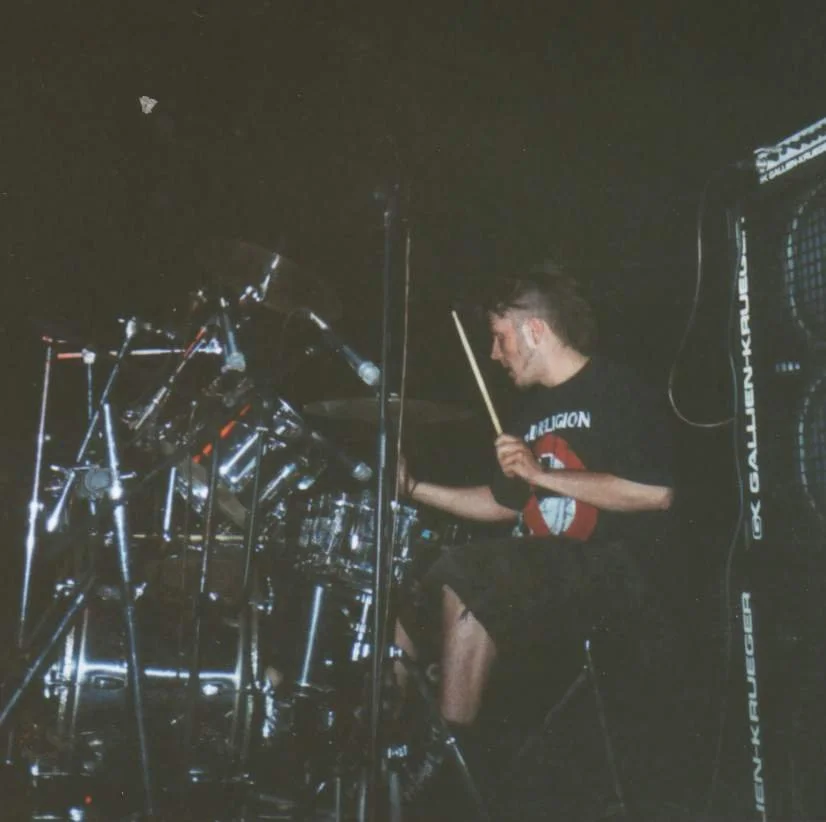 A young man playing a drum kit on stage, with dark background and a speaker beside him.