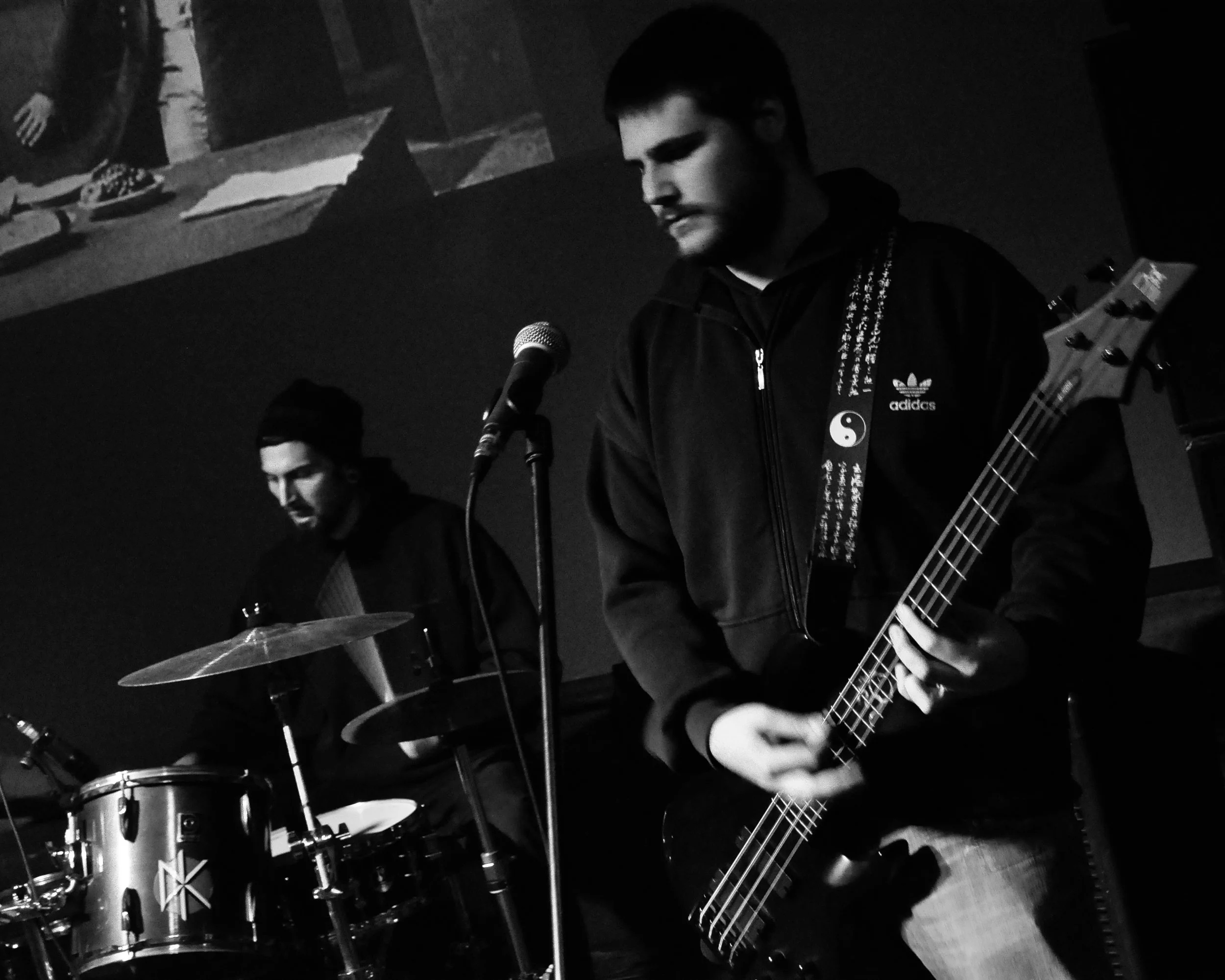 Black and white photo of two men performing music. One is playing drums and the other is playing an electric guitar. The man with the guitar has a beard and is wearing a hoodie with an Adidas logo and a yin-yang emblem. They are on a stage with a dar