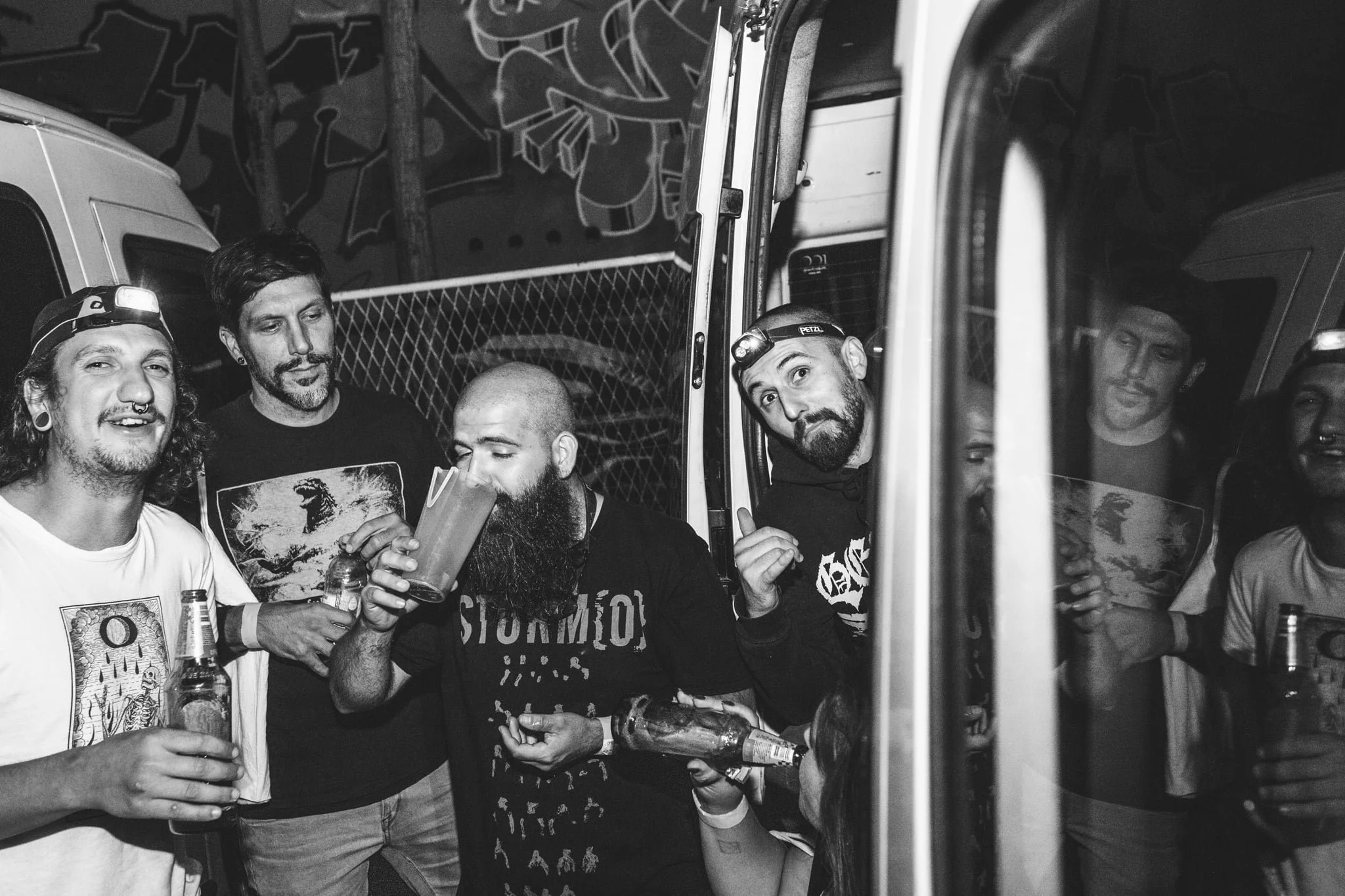 Group of young men socializing at a party, holding drinks, in a dimly lit space with graffiti on the wall.